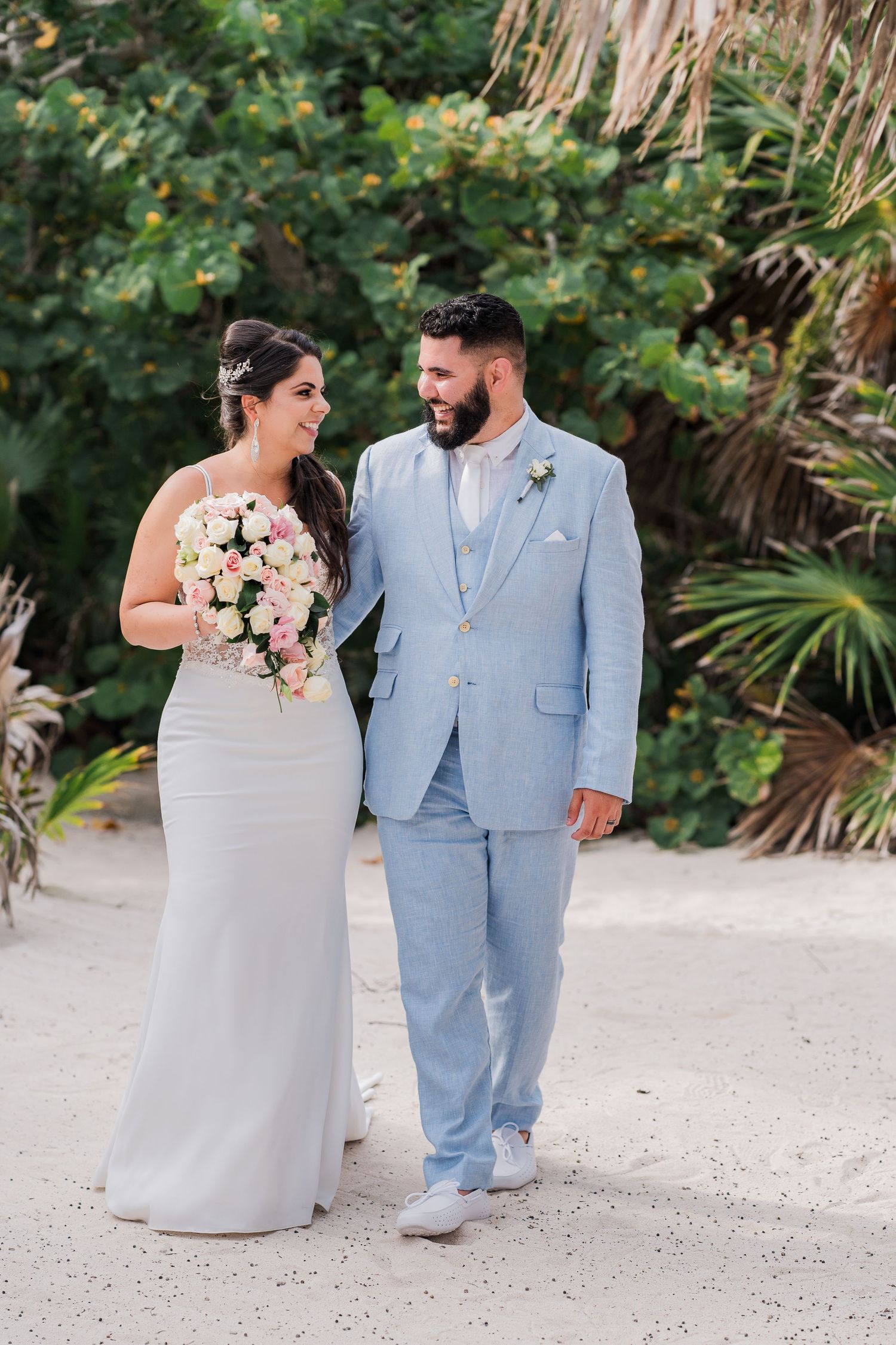 Bride in white gown holding pink bouquet walks with bearded groom in light blue suit on sandy beach.