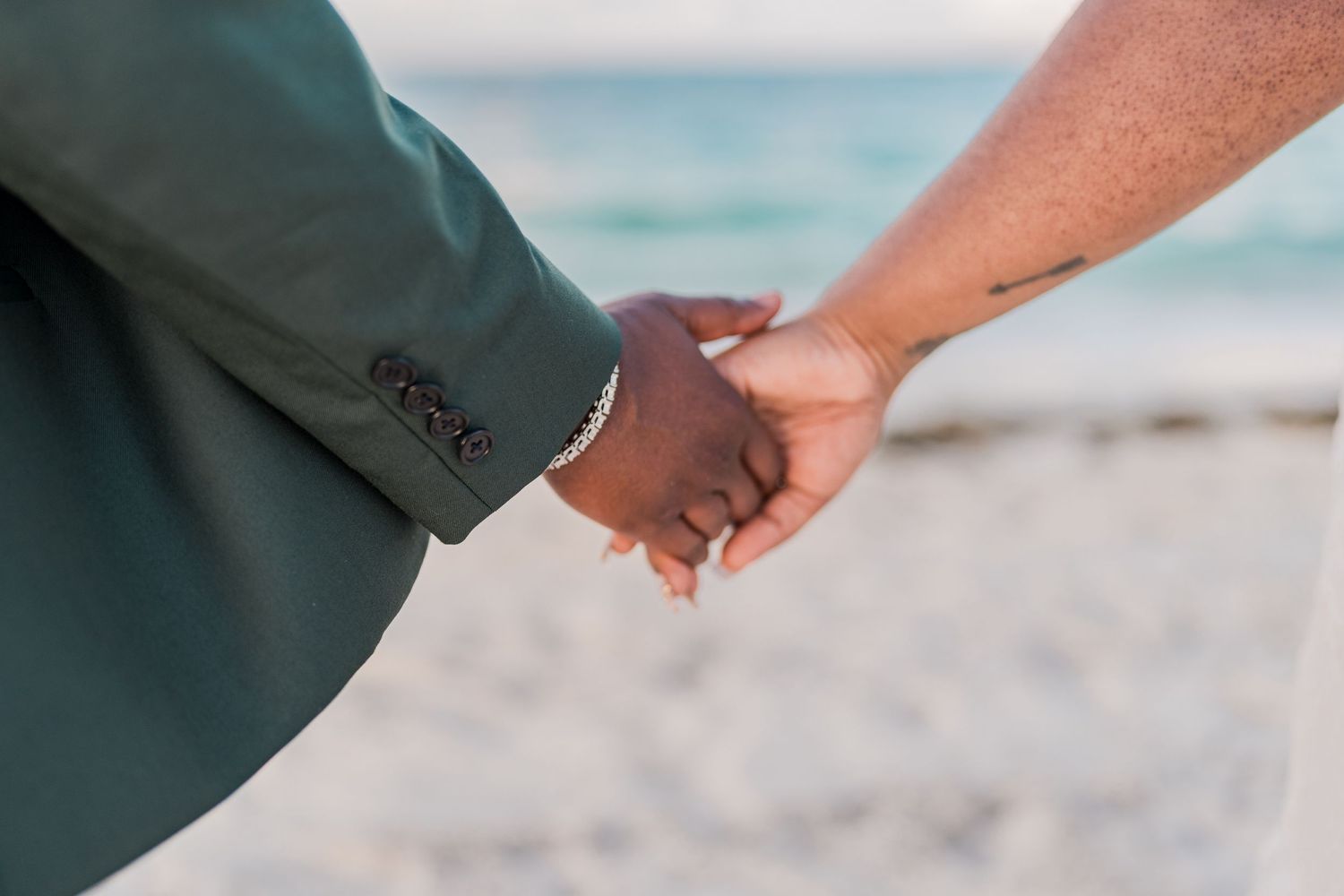 Close-up of interracial newlywed couple holding hands on a sandy beach with ocean in the background.