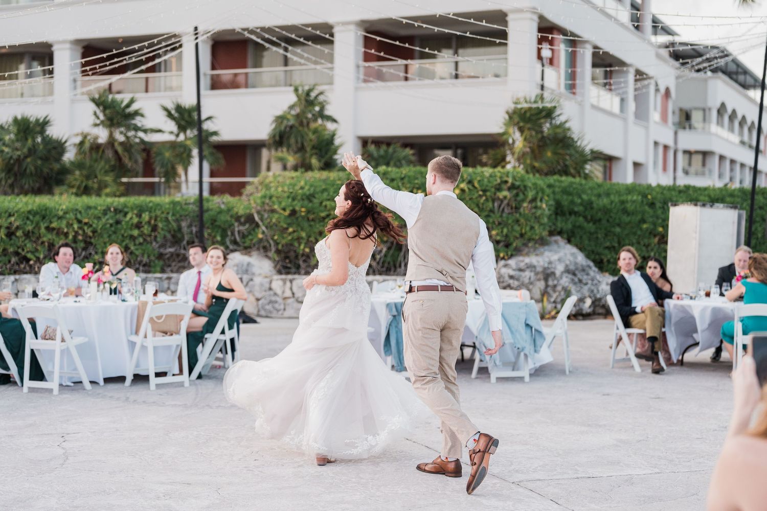 Groom twirls his bride in a white lace gown during their first dance at an outdoor destination wedding.