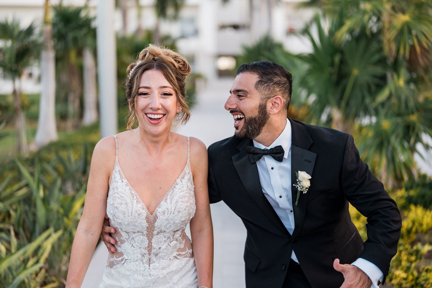 Laughing bride and groom walk together outdoors, bride in beaded V-neck gown, groom in black tuxedo.