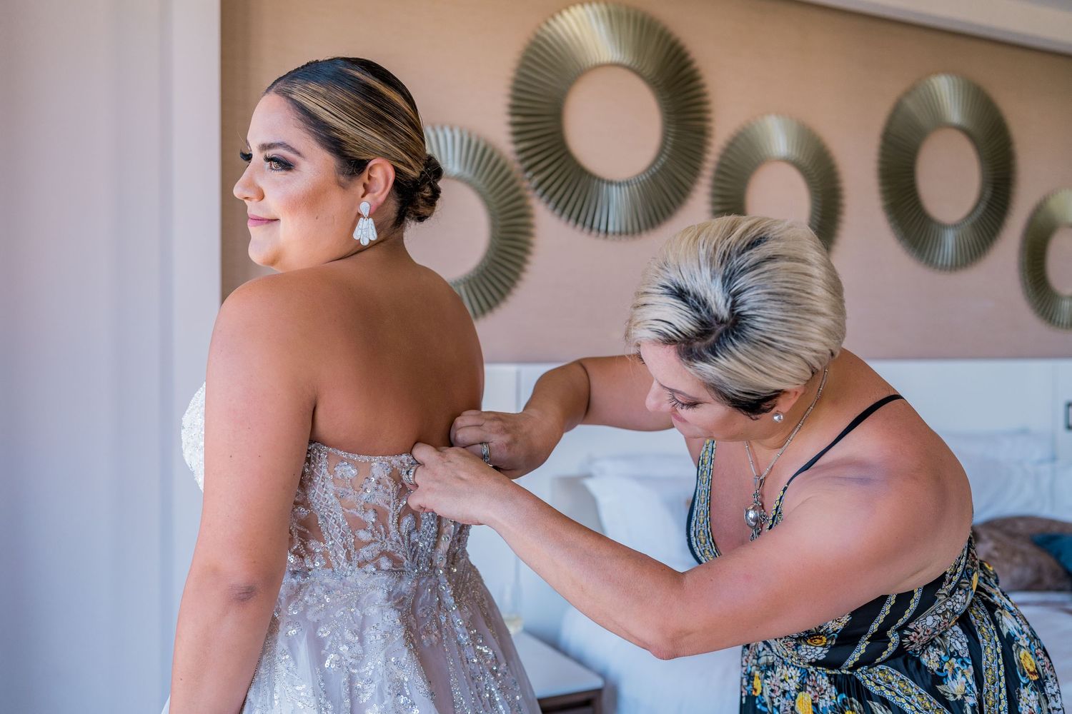 Mother helping bride fasten her strapless lace wedding gown in a hotel room before the ceremony.