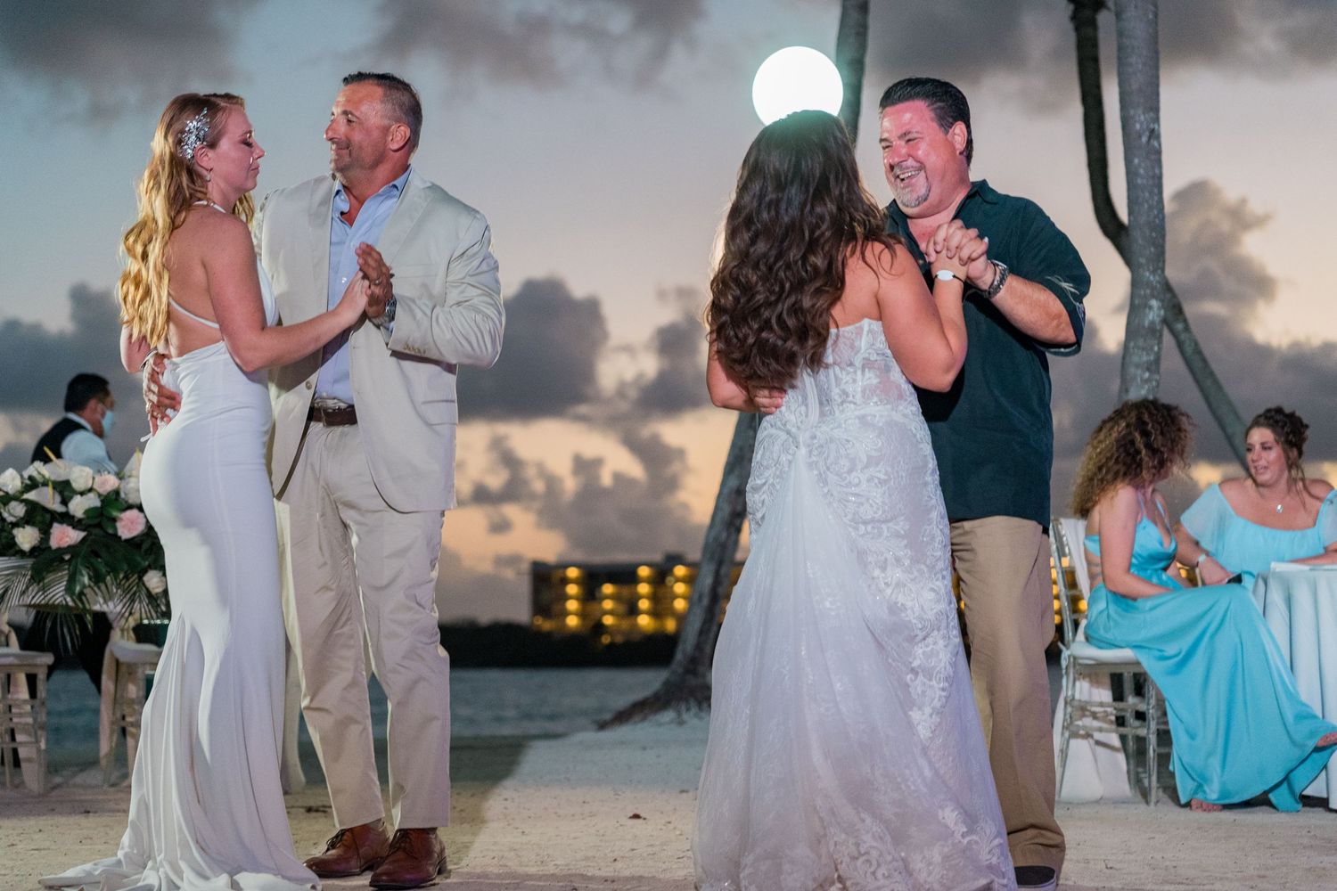 Two brides share father-daughter dances on tropical beach wedding reception at twilight.