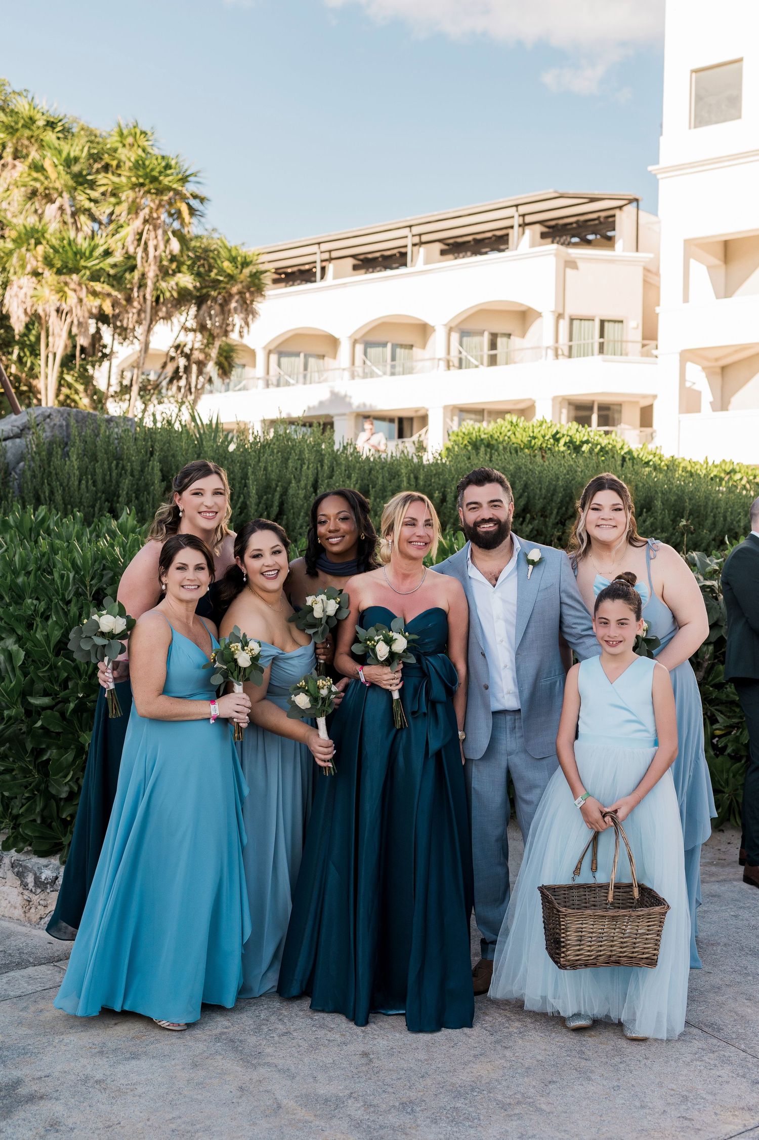Wedding party in blue gowns and gray suit poses outdoors at a resort, with flower girl holding a wicker basket.