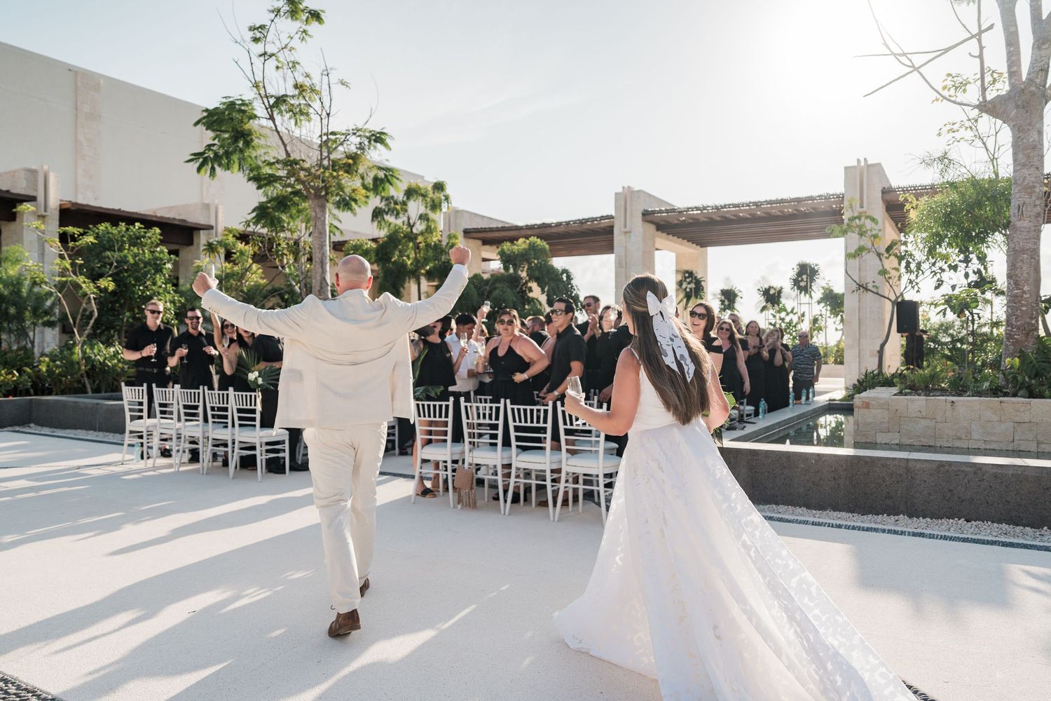 Bride and groom celebrate their outdoor wedding ceremony, raising hands joyfully as guests cheer in elegant white chairs.