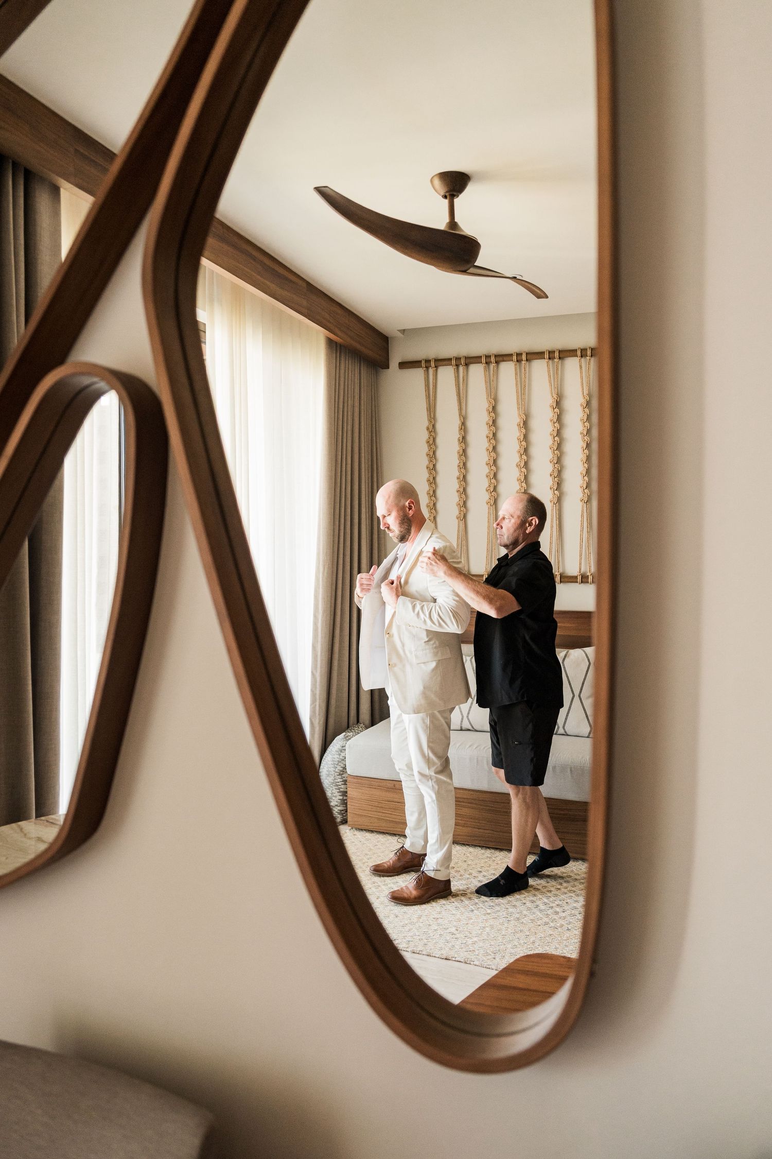 Two men in a hotel bedroom reflected in an ornate wooden mirror, one helping the other button his white shirt.