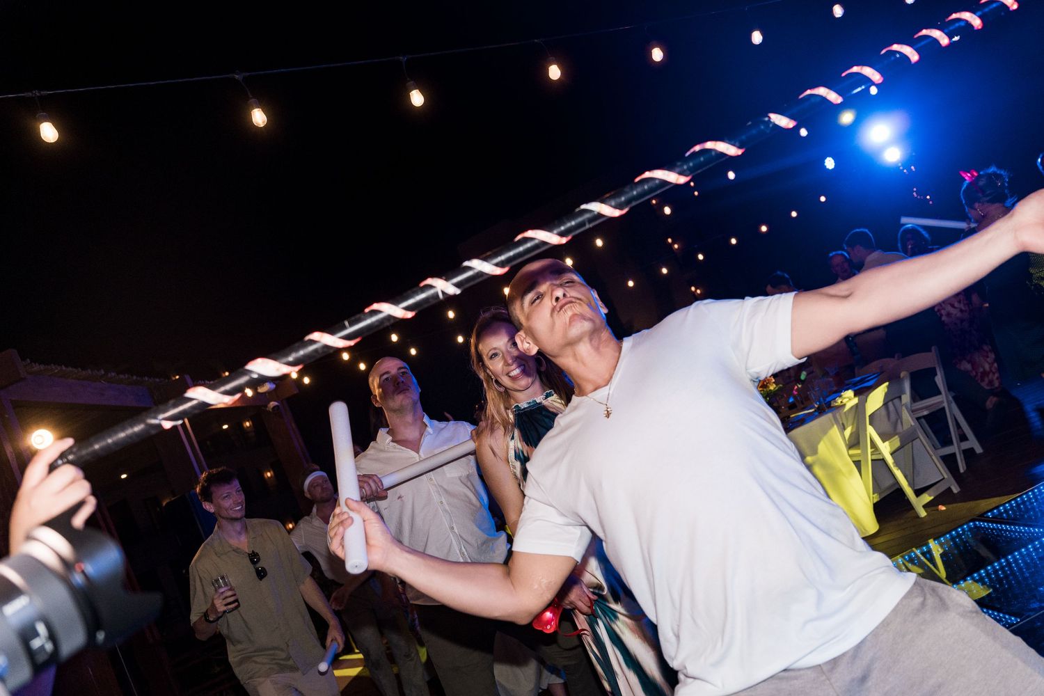 Man in white shirt dancing energetically at an outdoor night party with string lights and crowd behind him.
