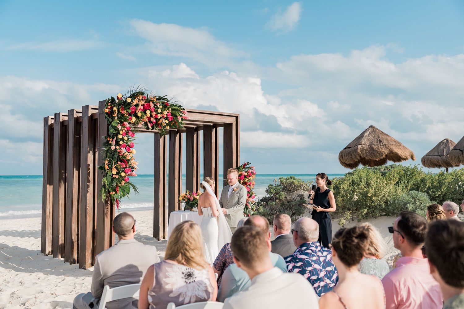 Outdoor beach wedding ceremony with floral arch, guests seated on white chairs, ocean backdrop, and thatched umbrellas under blue sky.