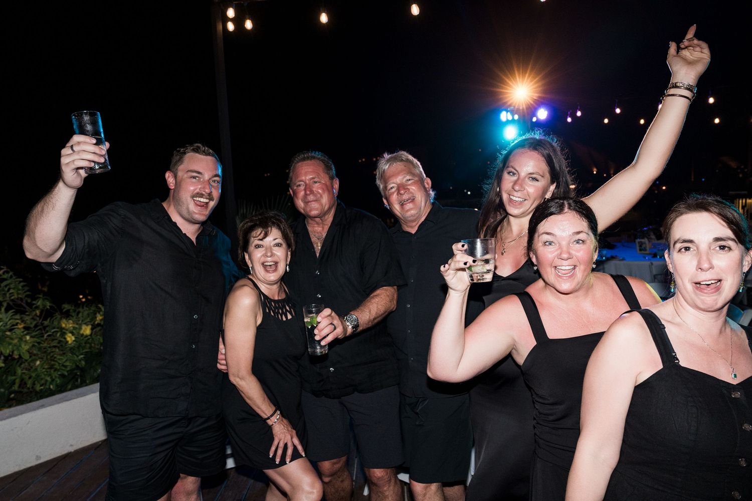 Group of friends in black outfits celebrating at a night party, raising drinks and smiling outdoors.