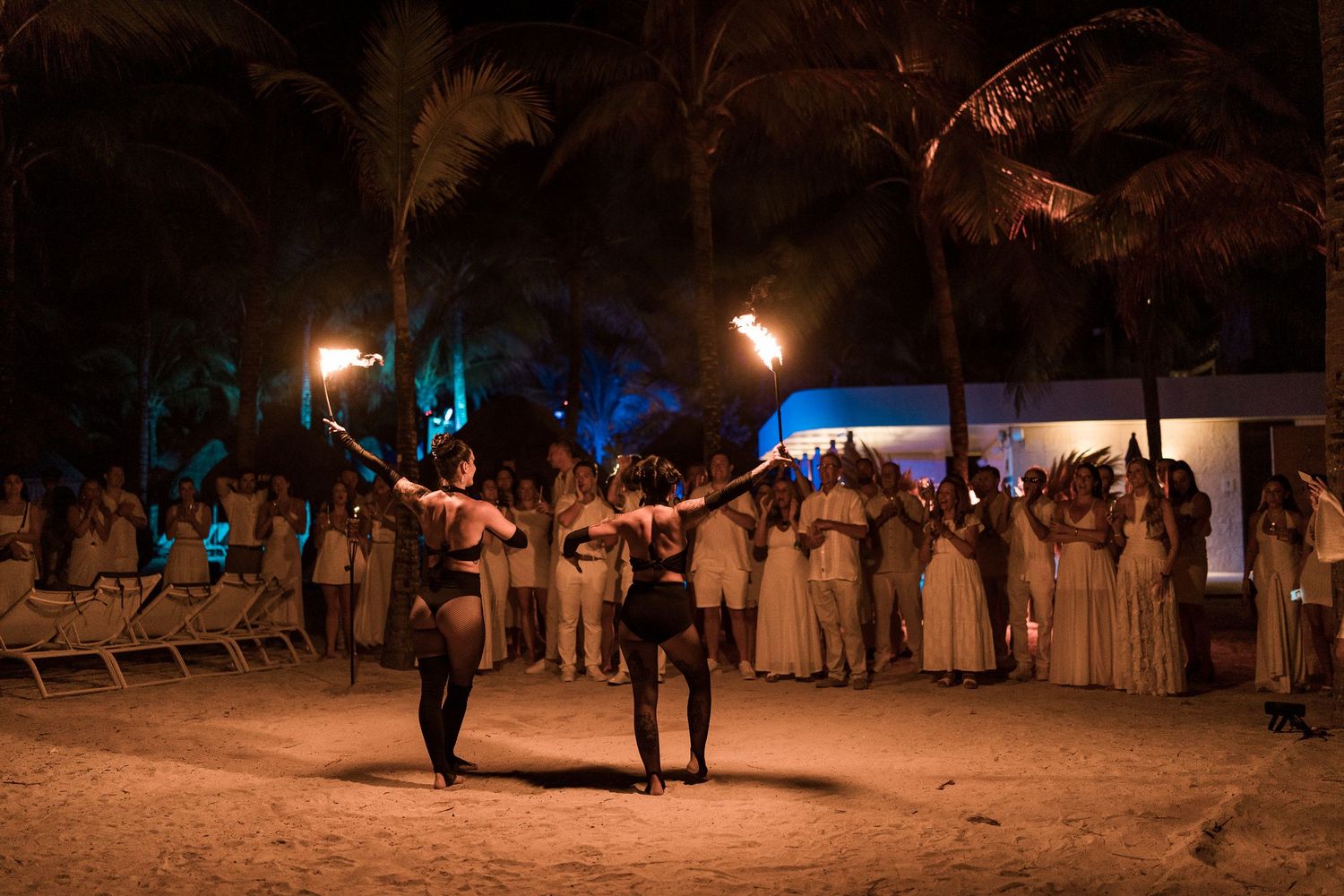 Fire dancers perform at a tropical beach luau at night with palm trees and white-clad audience watching.