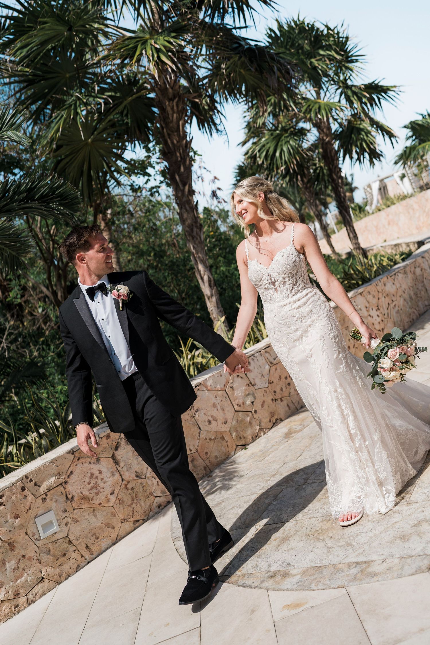 A bride and groom walk hand-in-hand on a tropical terrace, she in a lace gown, he in a black tuxedo.