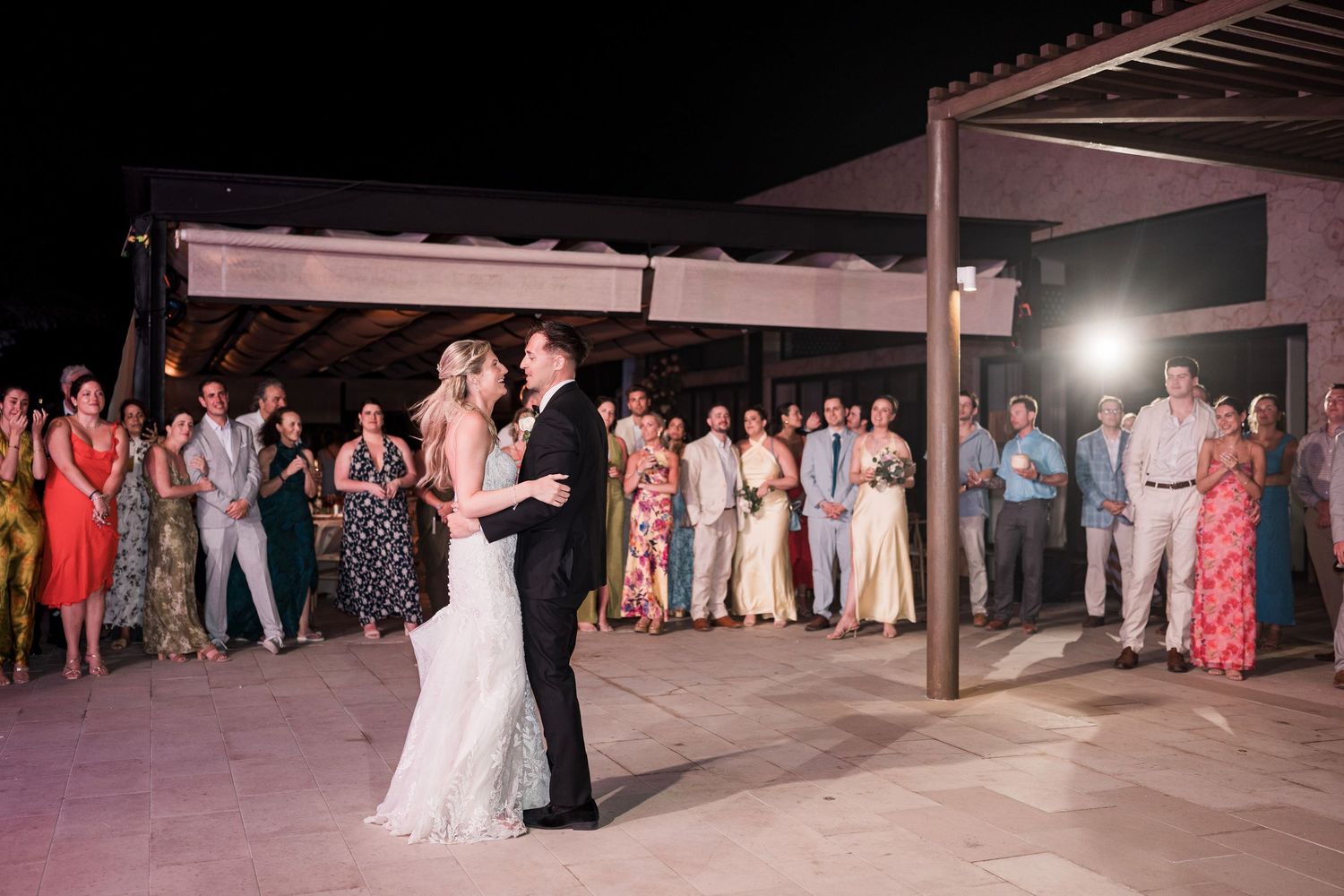 Bride and groom share first dance outdoors at night while wedding guests watch and celebrate around them.