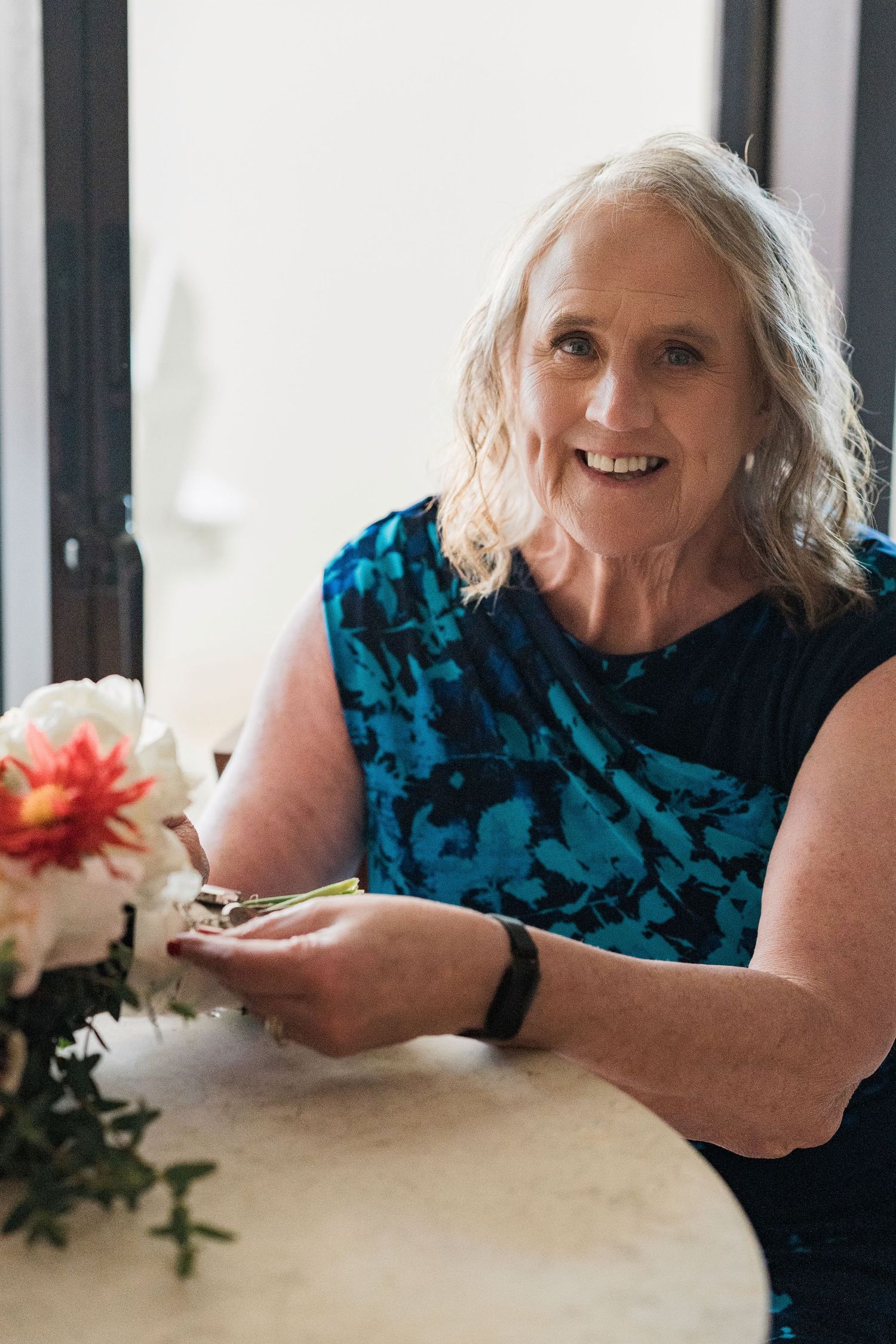 Smiling older woman with gray hair in blue top sitting at table with flowers.