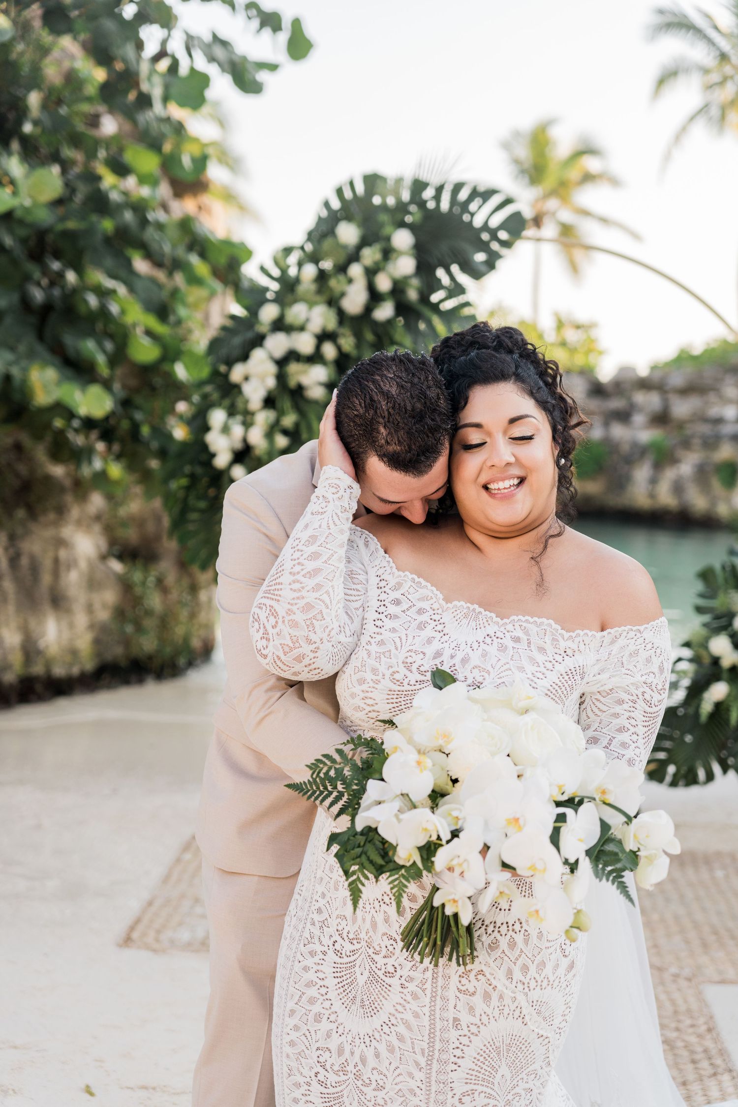Xcaret Arte Wedding - A bride in lace gown holds white bouquet as groom in tan suit nuzzles her neck at tropical wedding venue.