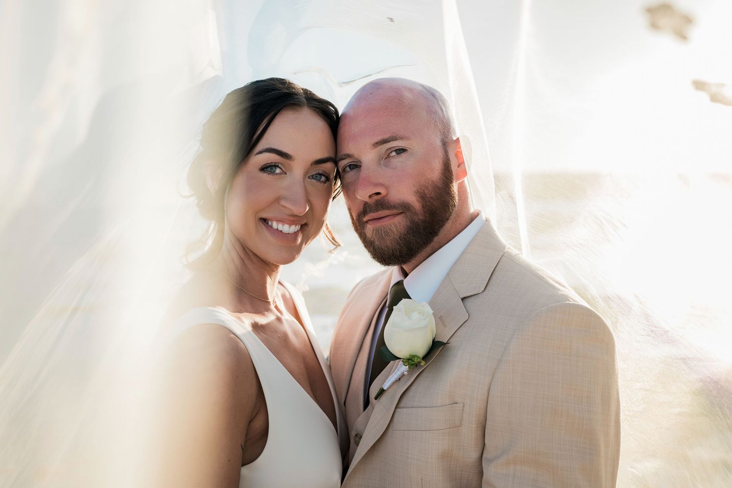 Bride and groom pose closely together under a flowing white veil, groom wearing a tan suit with a white rose boutonniere.