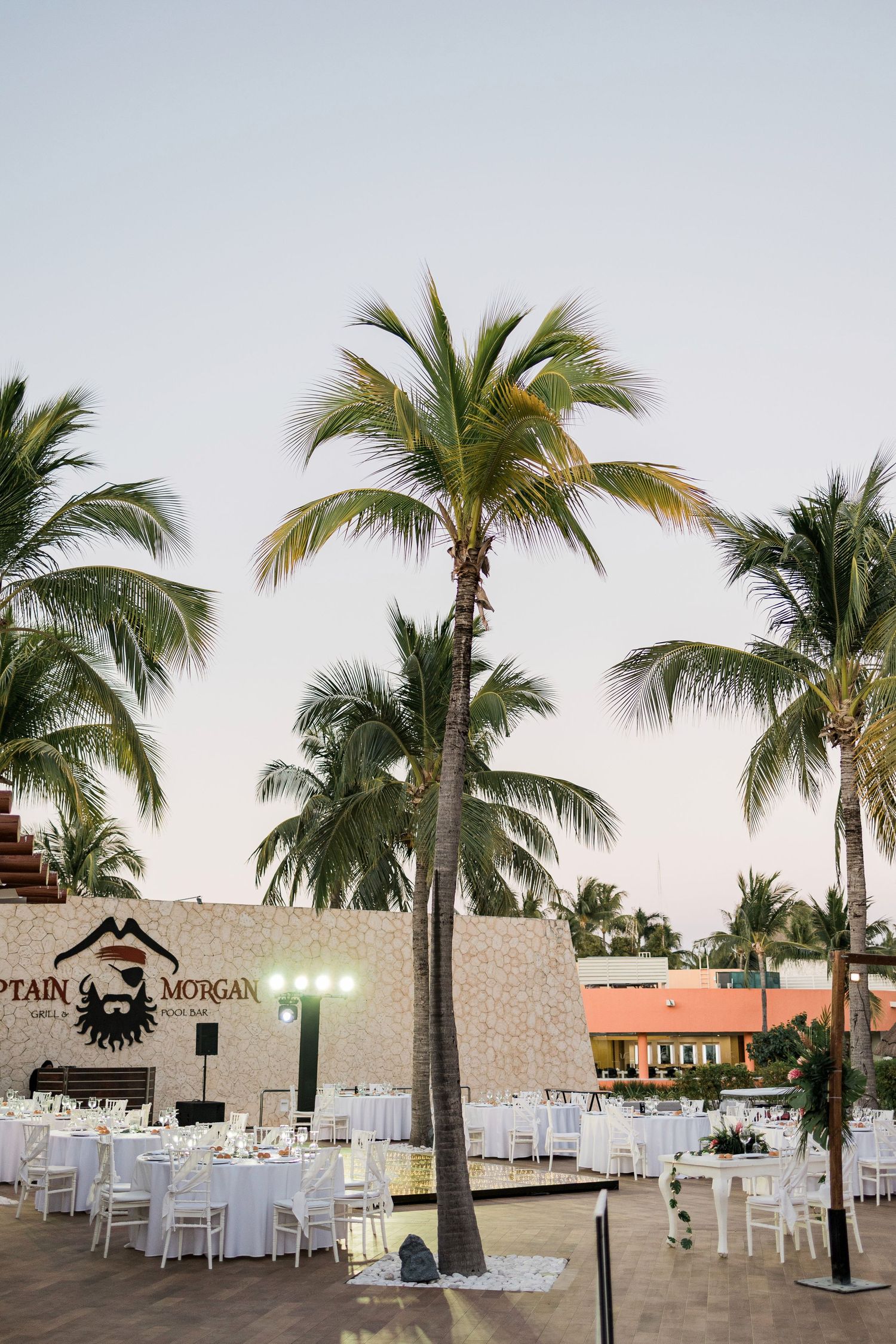 Outdoor wedding reception with white tables and chairs surrounded by palm trees near a Captain Morgan bar.