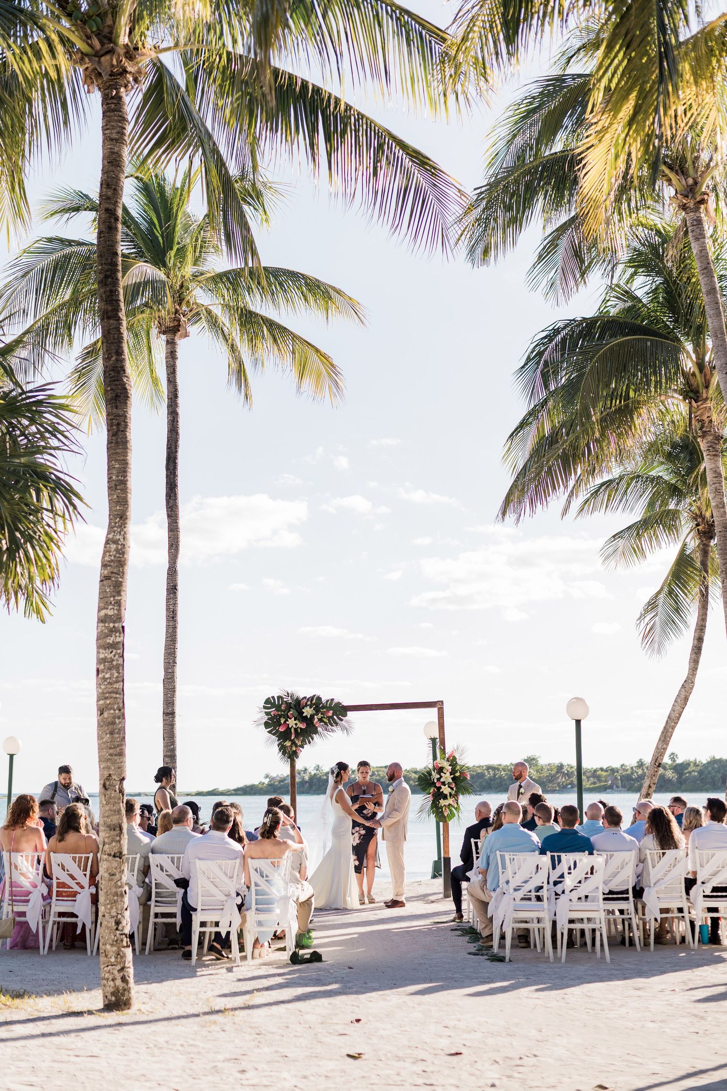 Outdoor beach wedding ceremony under palm trees by the water, with guests seated in white chairs around a floral arch.
