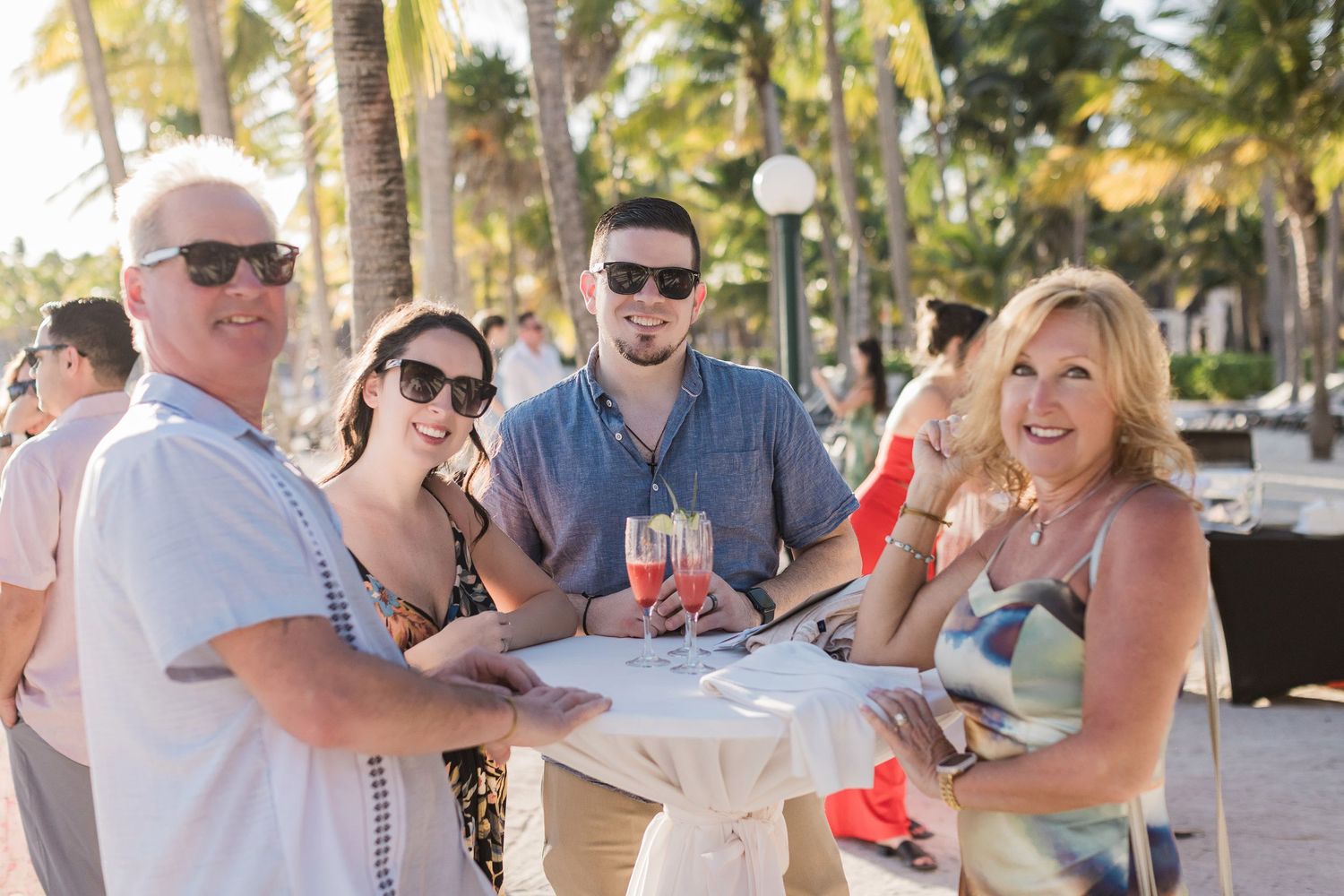Four people smiling and holding drinks at an outdoor tropical resort with palm trees in the background.