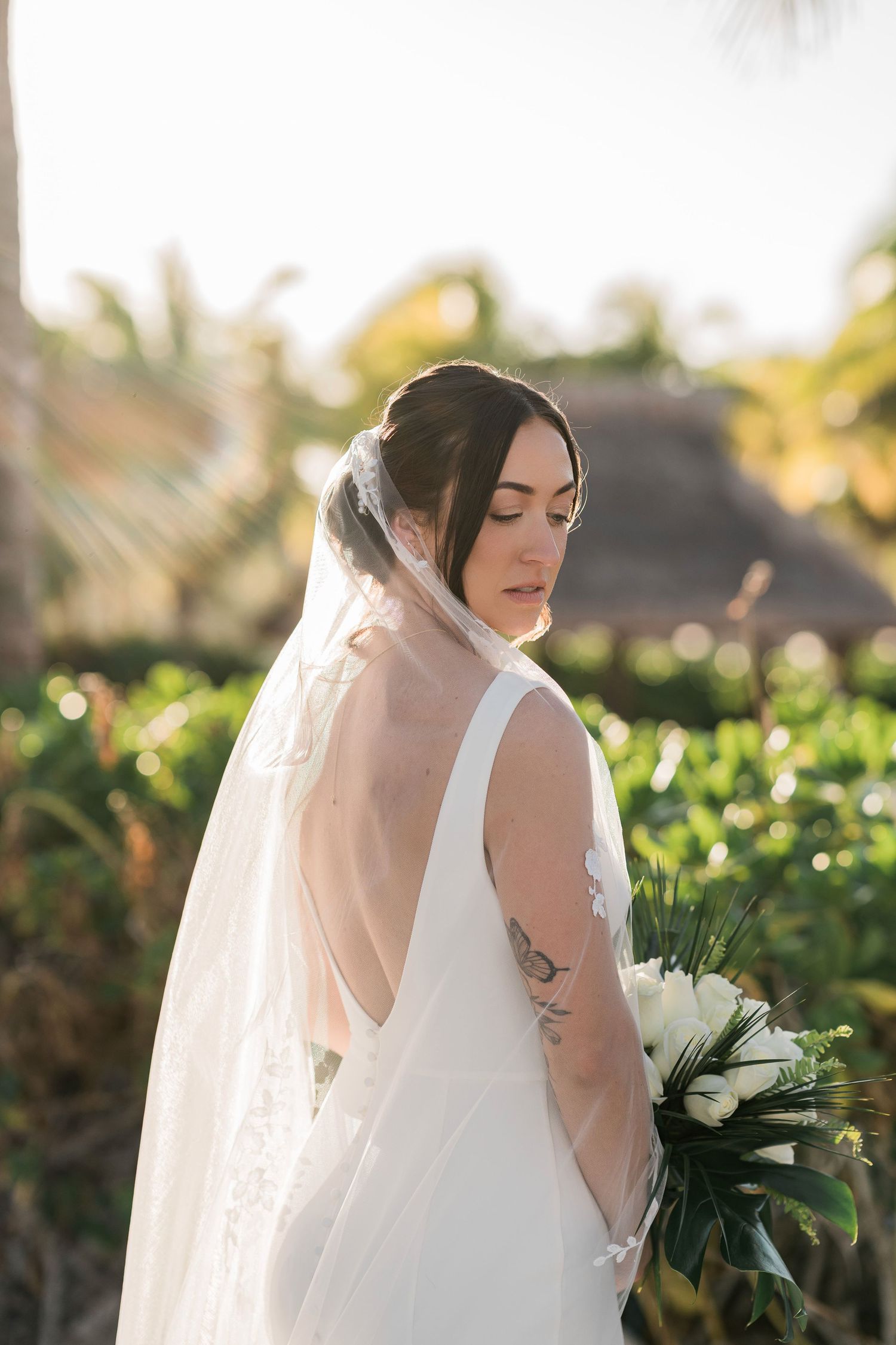 Bride in backless white gown and veil holds white bouquet, posing outdoors with tropical palms and golden sunlight.