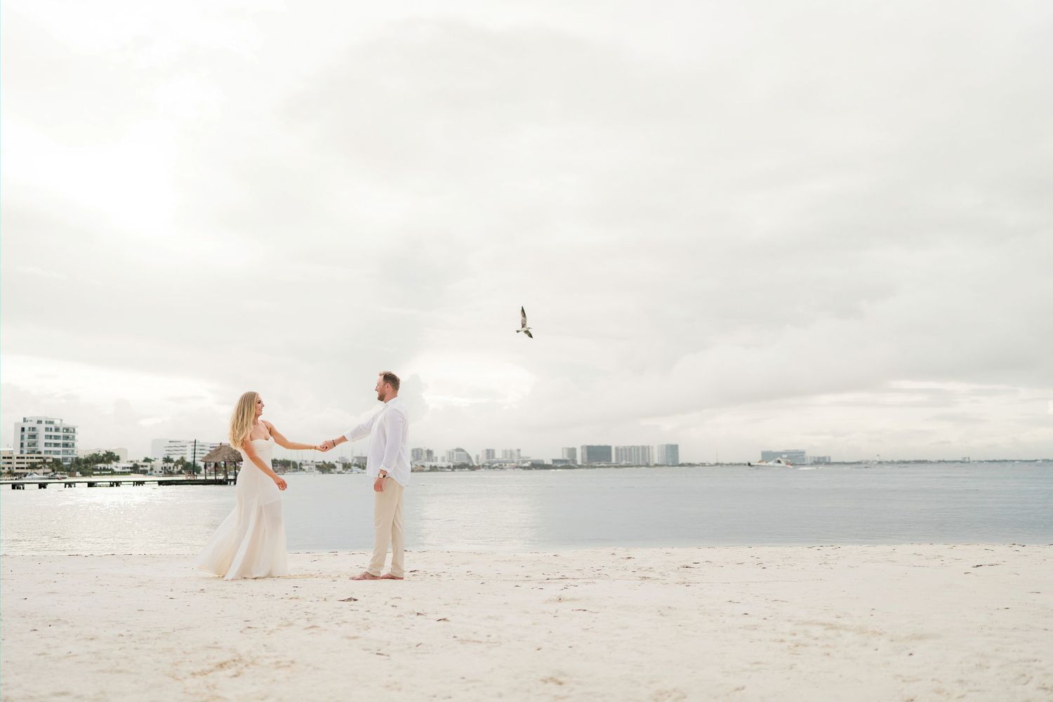 Cancun Engagement Photoshoot - Couple holds hands on a white sand beach with city skyline and calm water backdrop on a cloudy day.