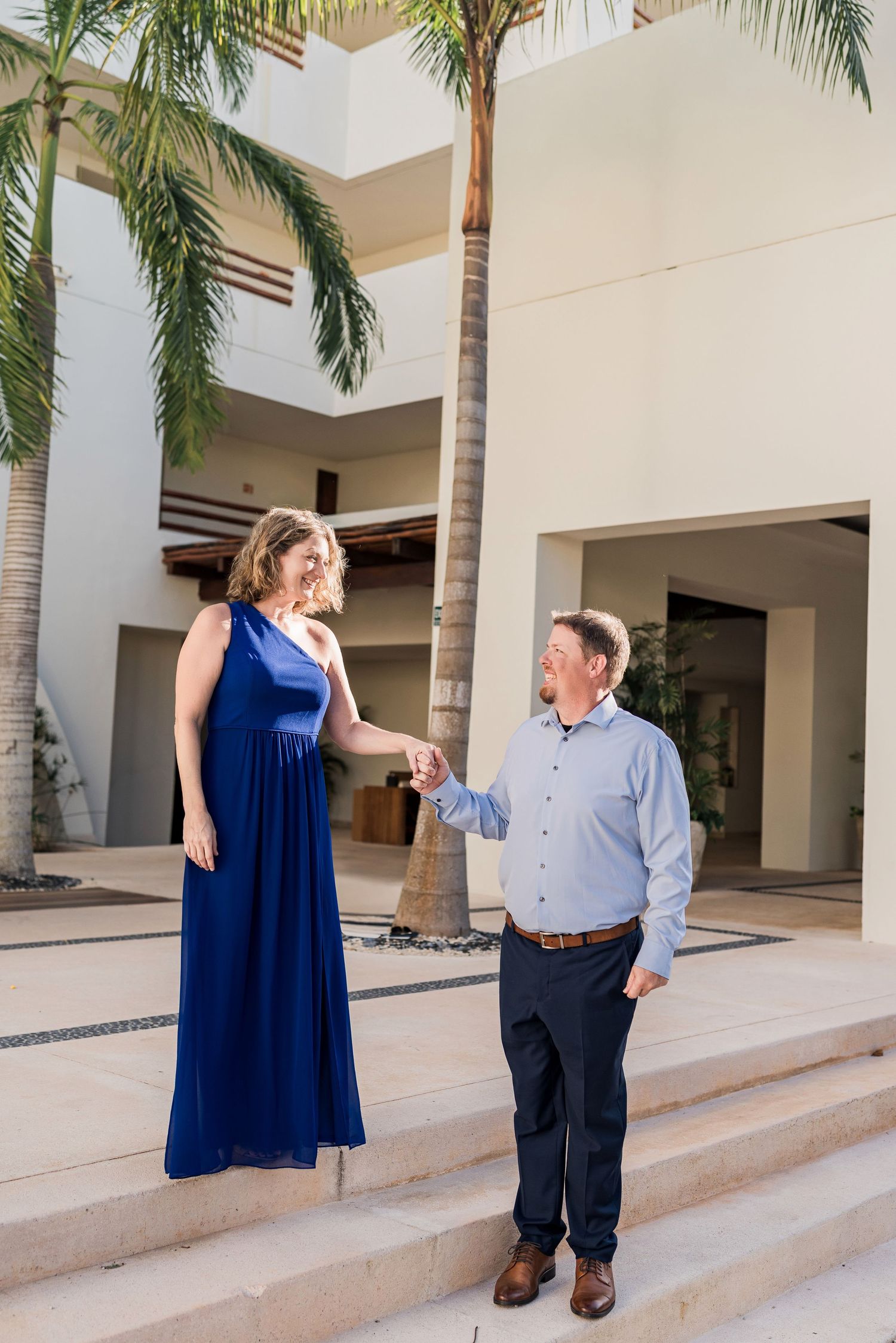 Secrets Akumal Wedding - Couple in elegant blue dress and blue shirt hold hands on resort steps with palm trees.