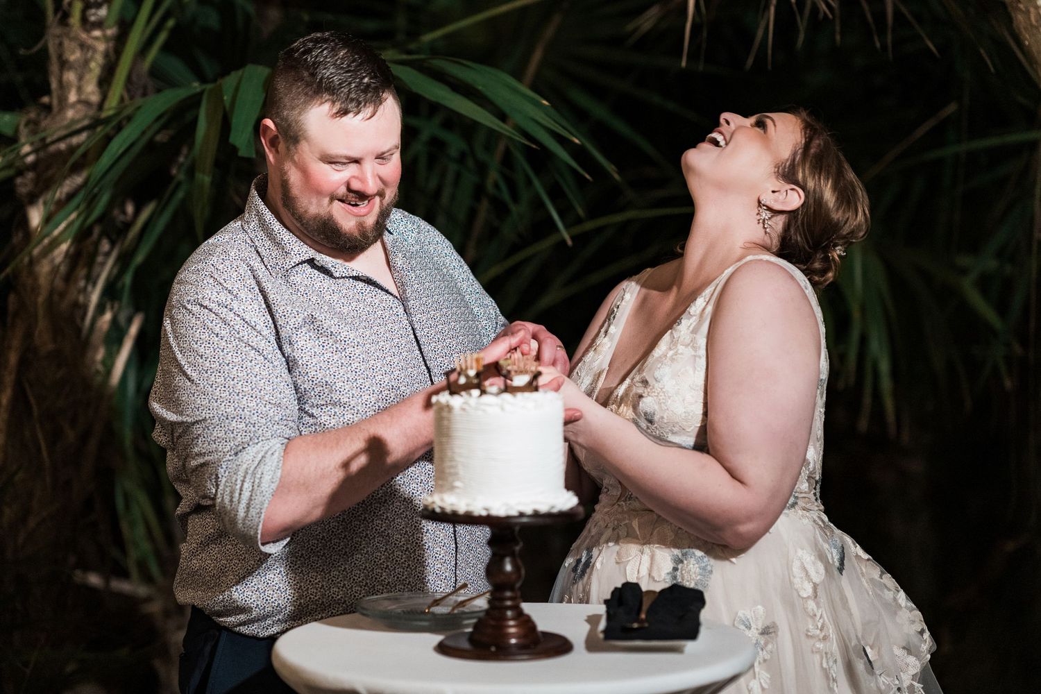 Secrets Akumal Wedding - Bride and groom laughing while cutting white wedding cake outdoors at night surrounded by tropical greenery.