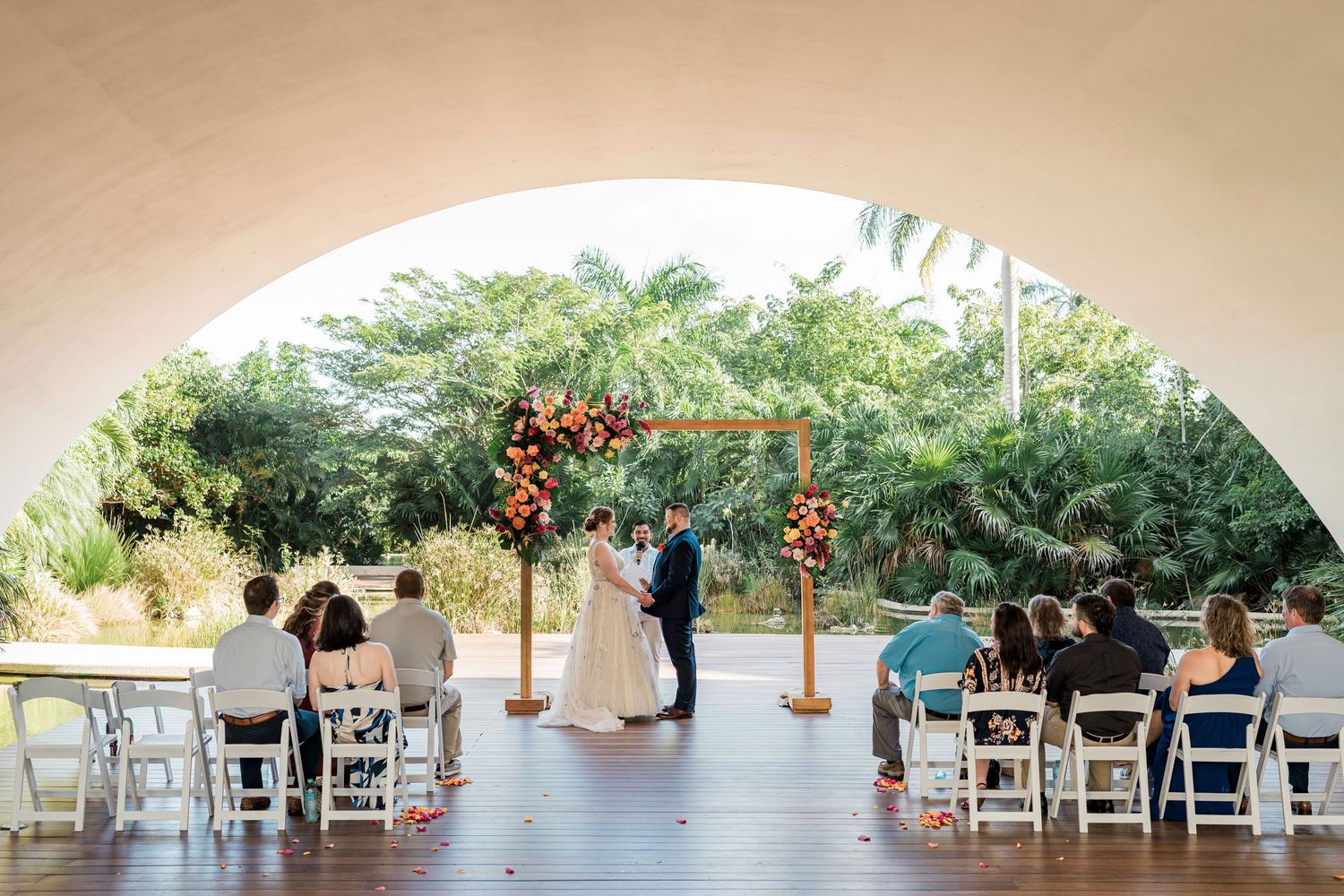 Secrets Akumal Wedding - Couple exchanges vows under floral arch at outdoor wedding ceremony with white chairs and lush greenery backdrop.