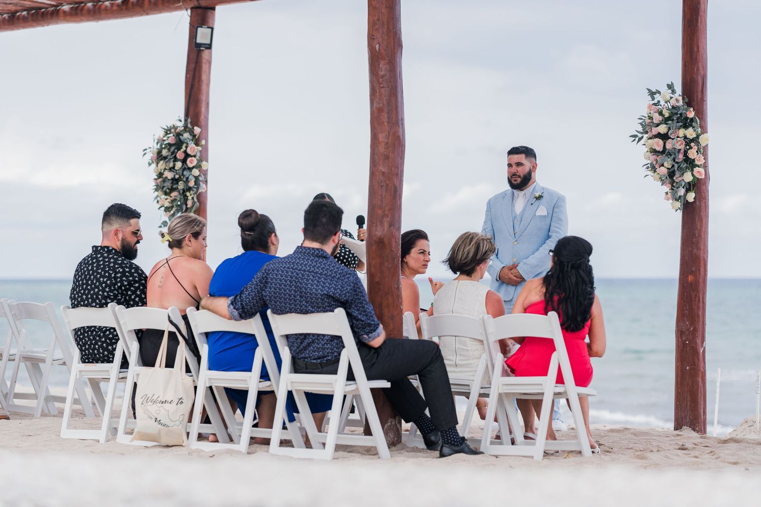 Small intimate beach wedding ceremony with guests seated in white chairs under wooden pergola.