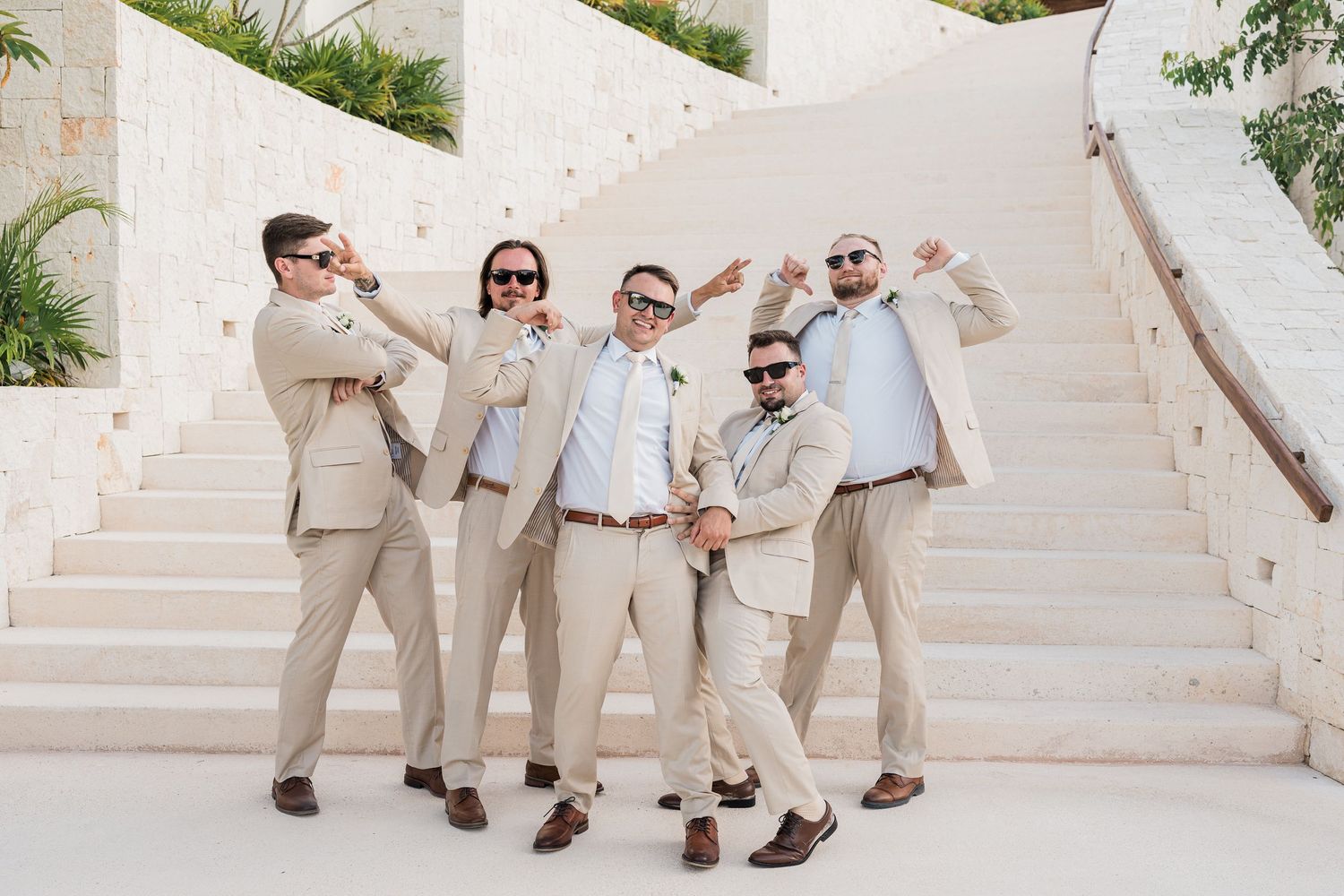 Five groomsmen in tan suits and sunglasses pose playfully on white stone steps at a destination wedding.