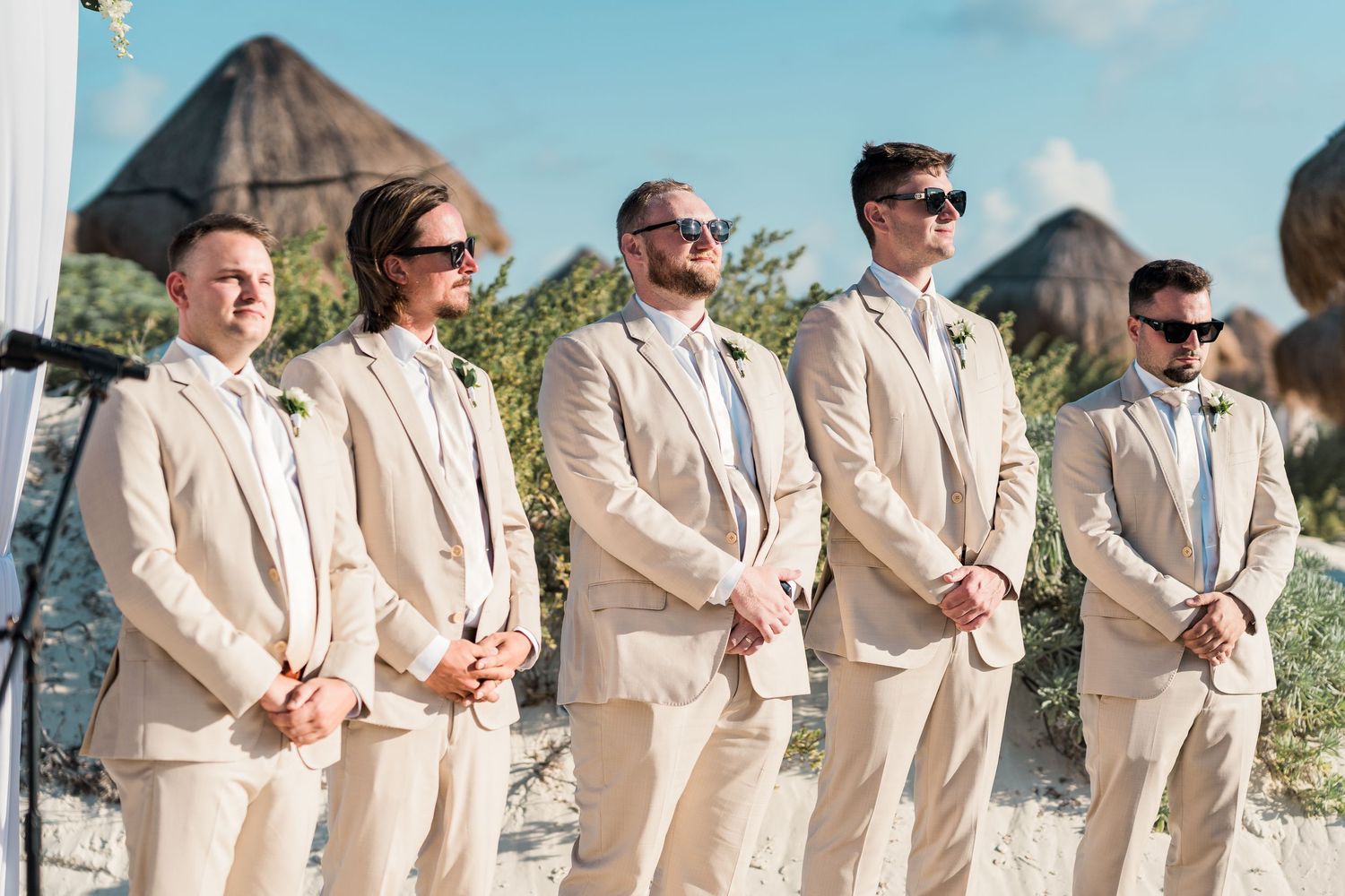Groom and groomsmen in matching tan suits wearing sunglasses at tropical beach wedding ceremony.