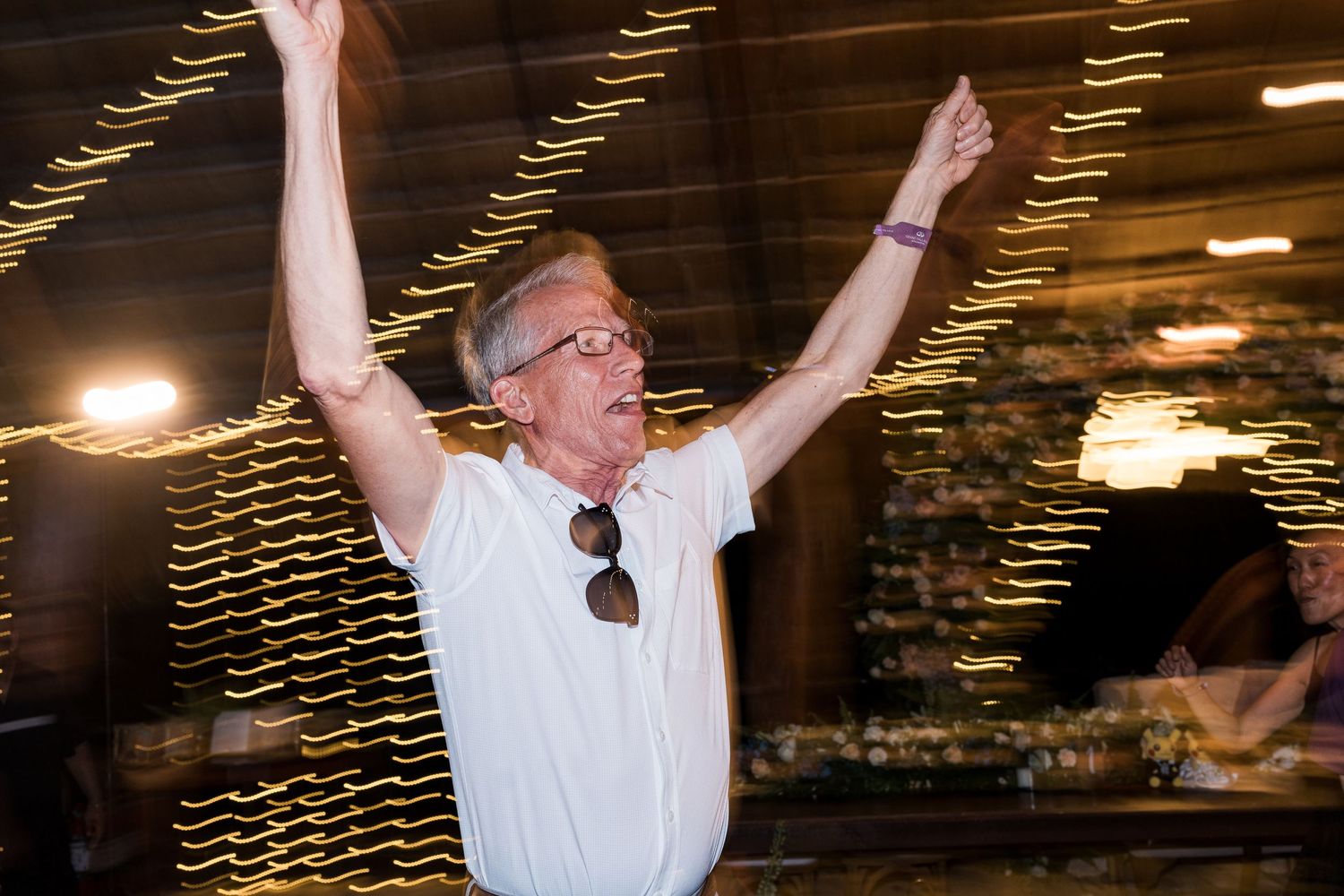 Joyful elderly man raises arms triumphantly on wedding reception dance floor under lights.