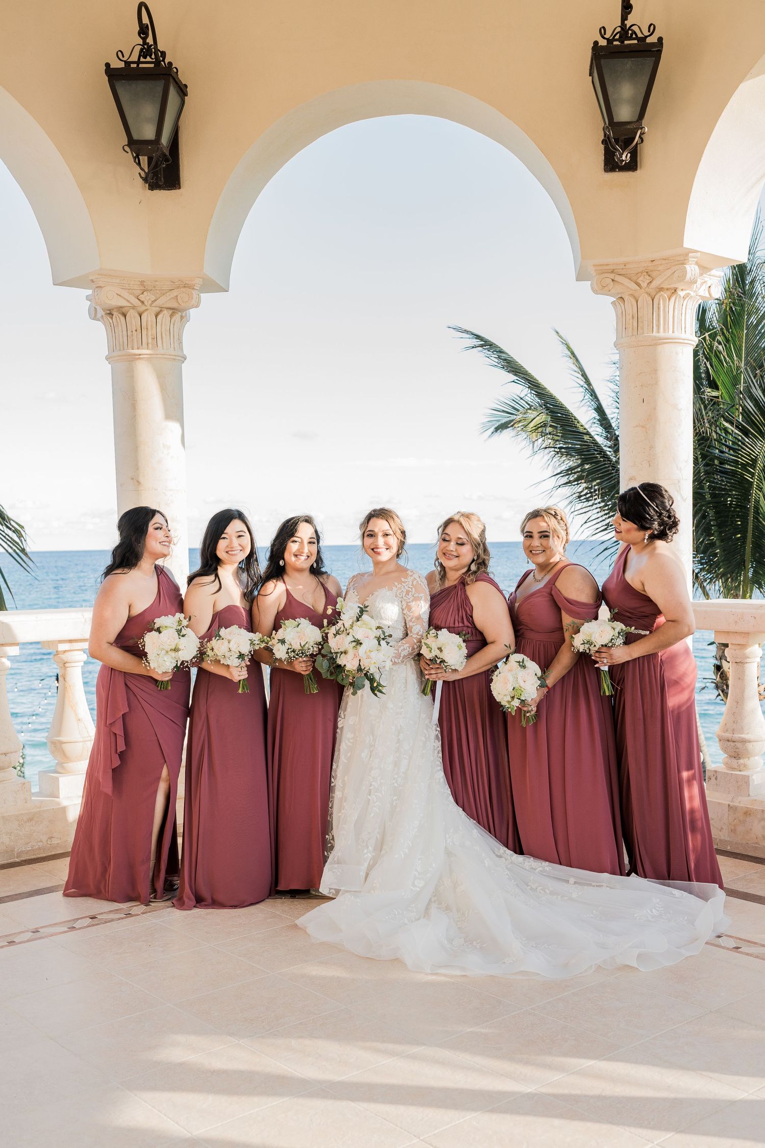 Bride with six bridesmaids in dusty rose gowns holding white bouquets at oceanfront wedding venue.