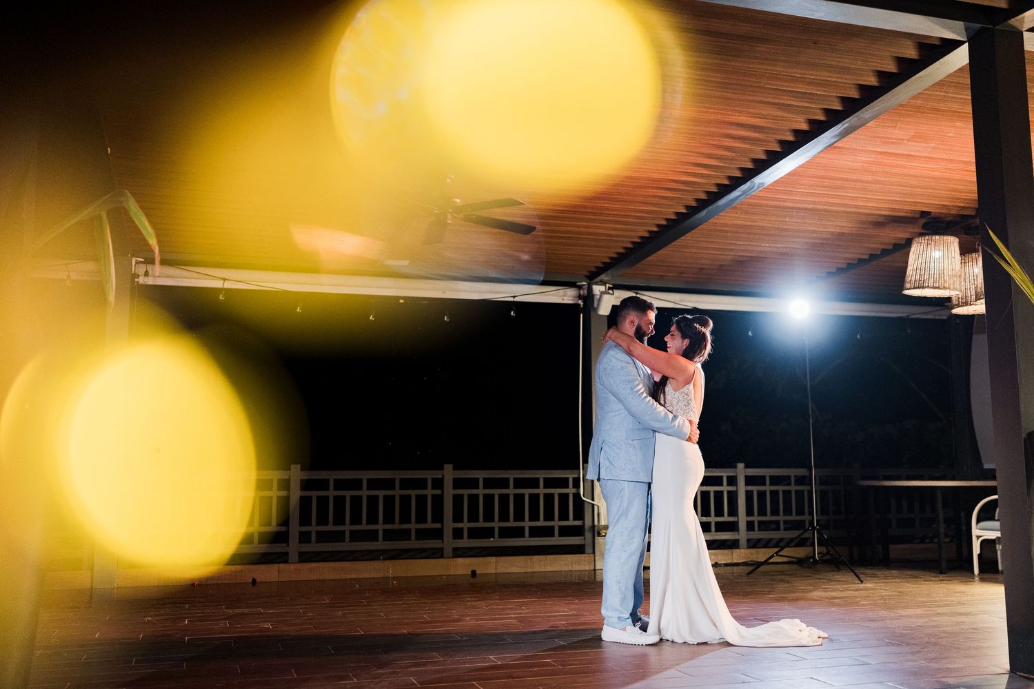 Bride and groom silhouetted in romantic embrace on outdoor dance floor under pergola with dramatic golden bokeh lighting.