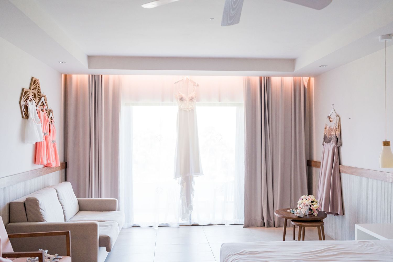 Elegant bridal suite with wedding gown hanging by sheer curtains, pink bridesmaid dresses, and soft natural light.