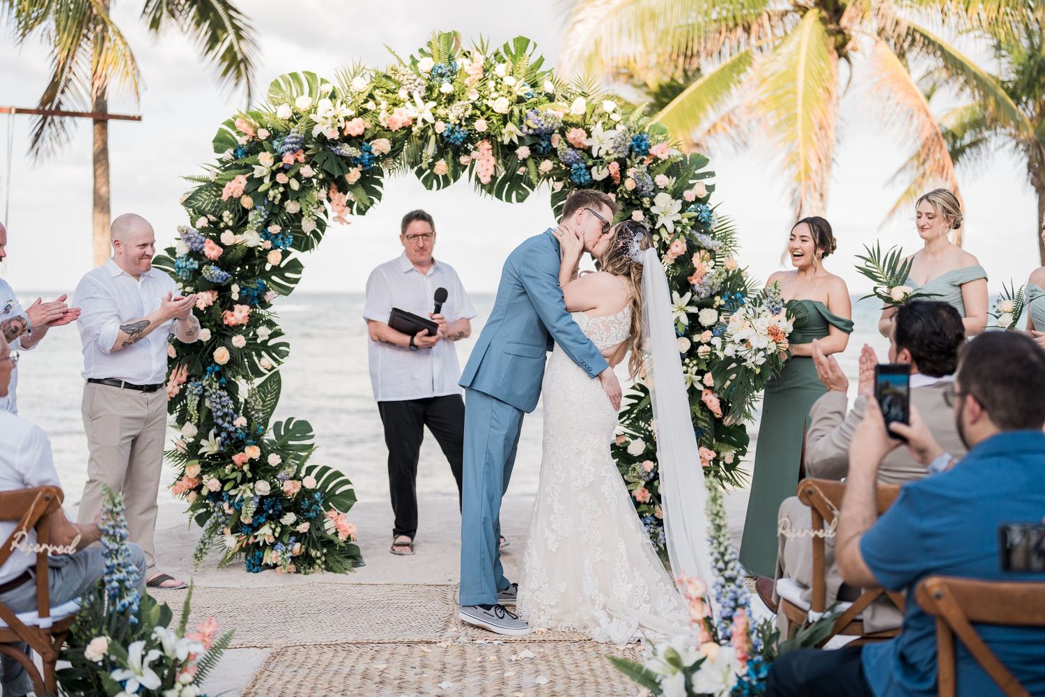 Bride and groom share first kiss under floral arch at tropical beach wedding ceremony.