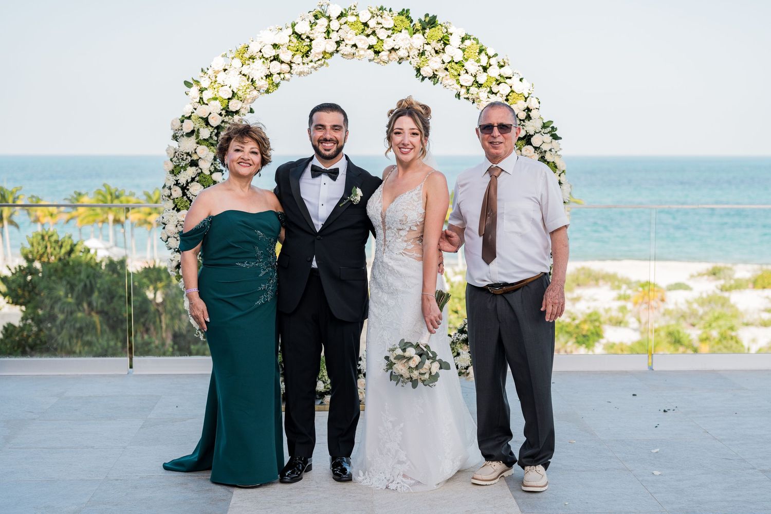 Newlyweds pose with parents under white rose arch at scenic oceanfront wedding ceremony.