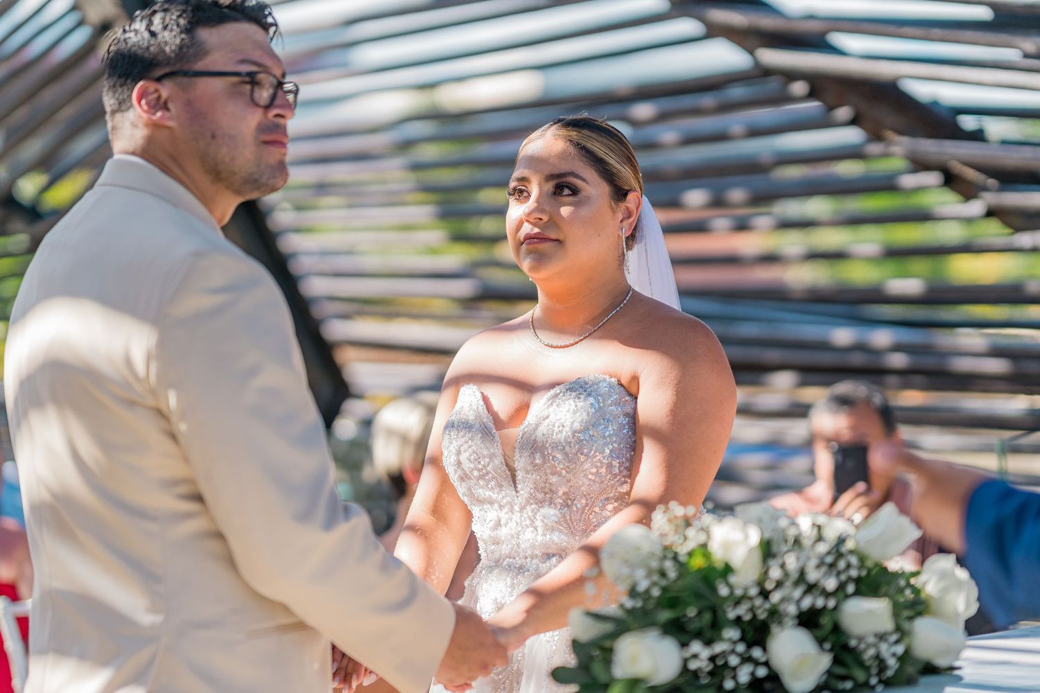 Bride in beaded gown and veil gazes at groom during emotional outdoor wedding ceremony.