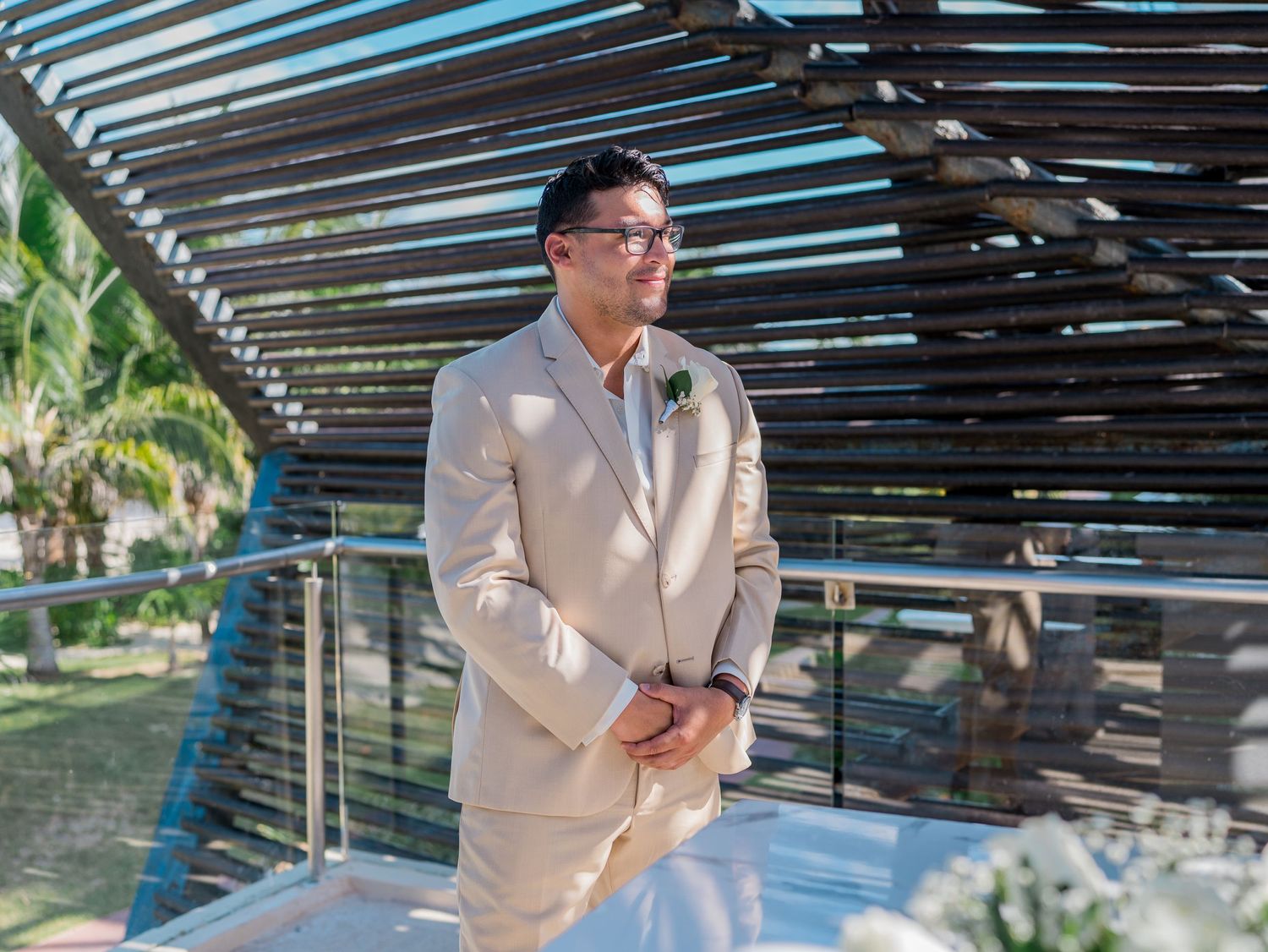 Groom in beige suit with boutonniere stands at outdoor wedding ceremony venue with palm trees.