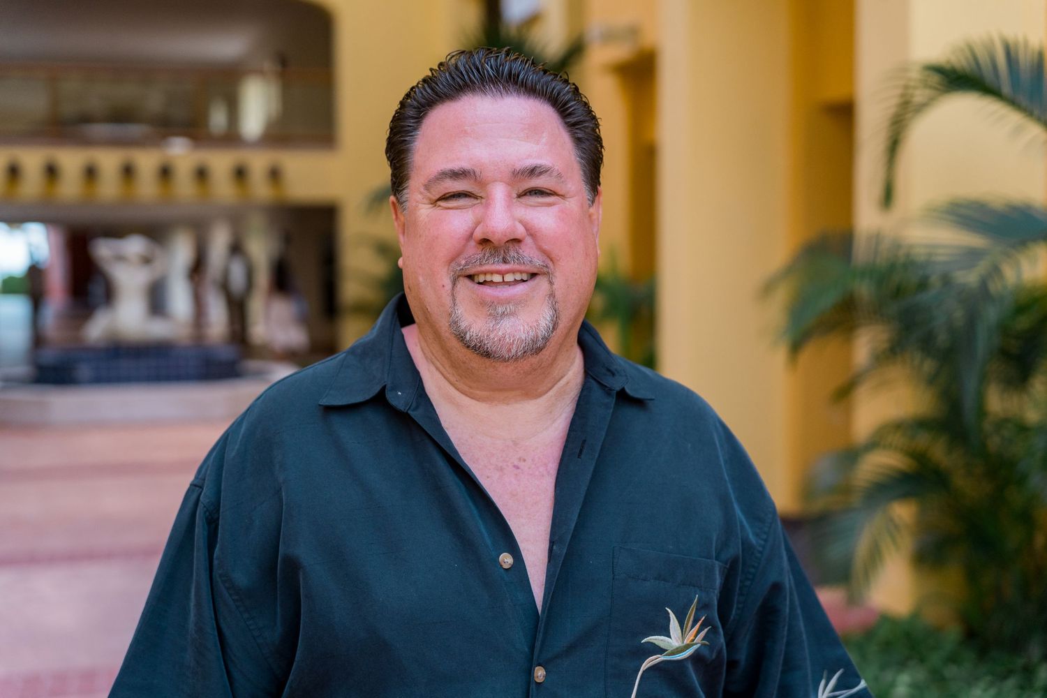 Smiling man in a dark green floral shirt posing outdoors at a tropical resort venue.