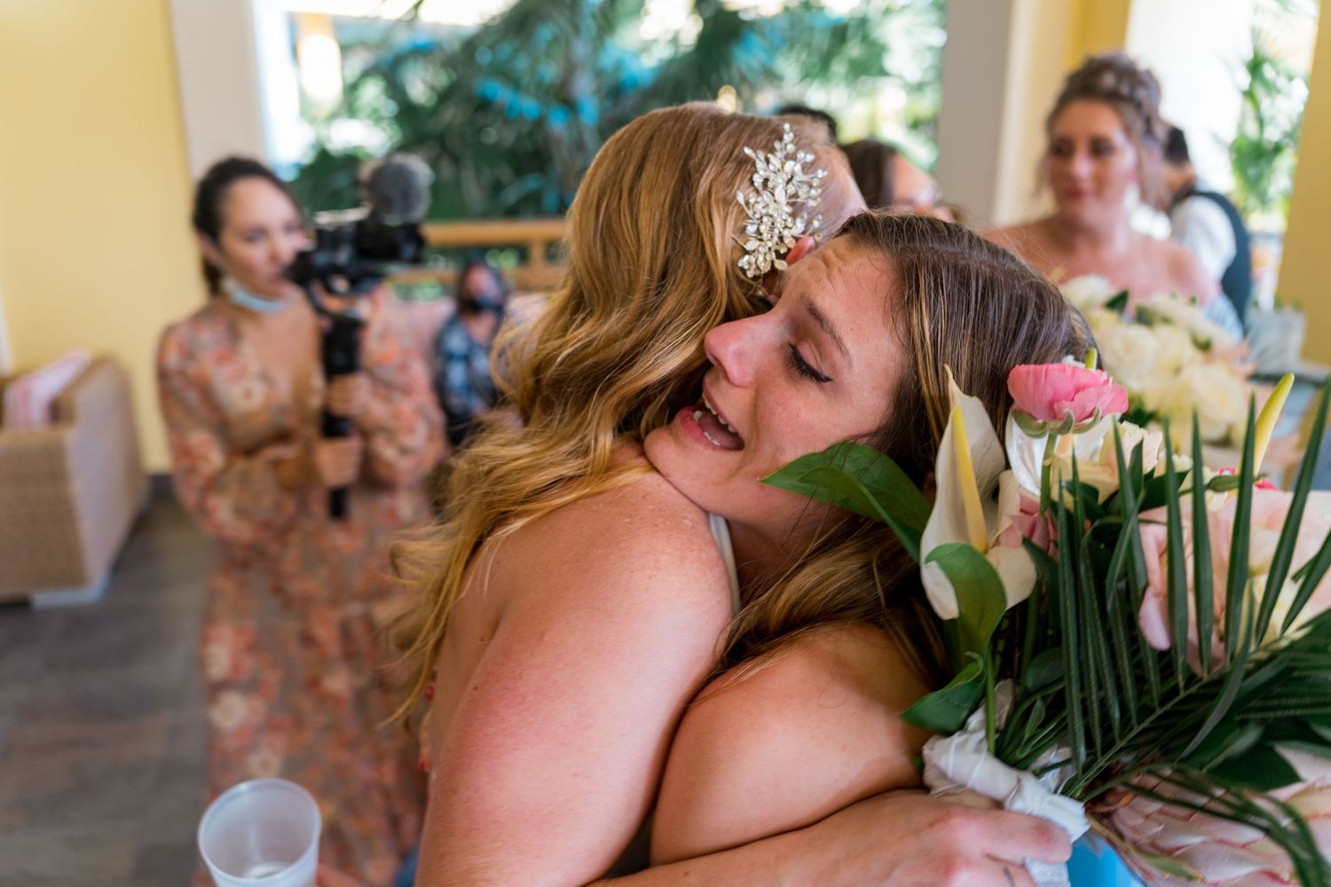 Two women sharing a warm embrace at a wedding, one holding a bouquet of pink flowers.