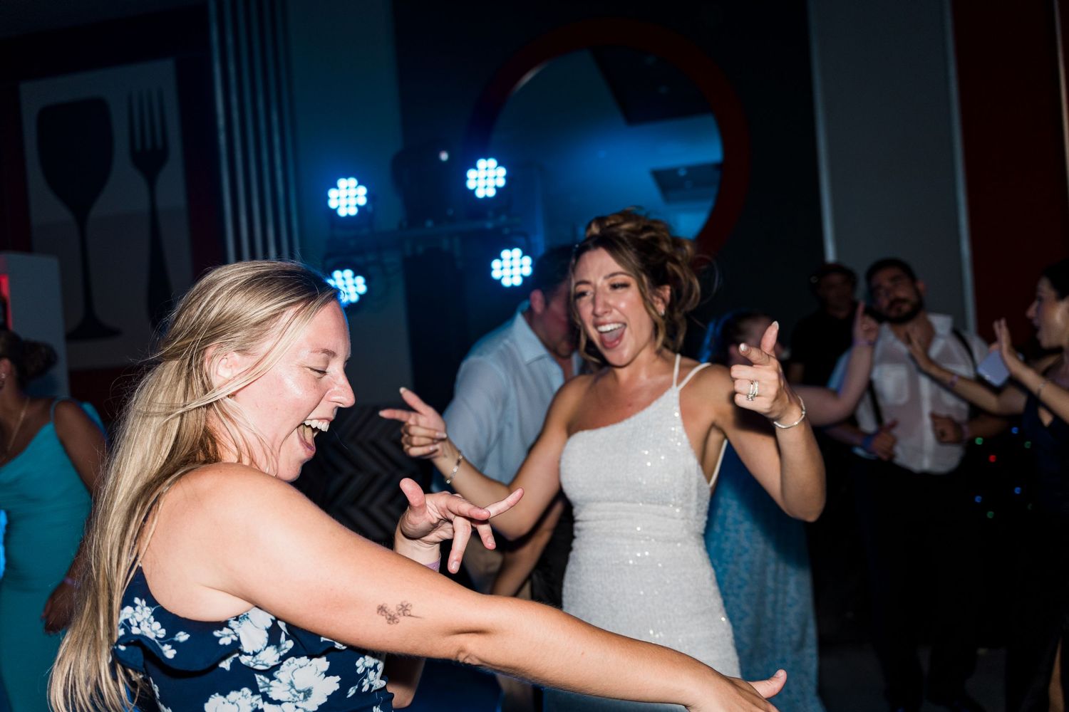 Bride and guest dancing joyfully at wedding reception with DJ lights in background.