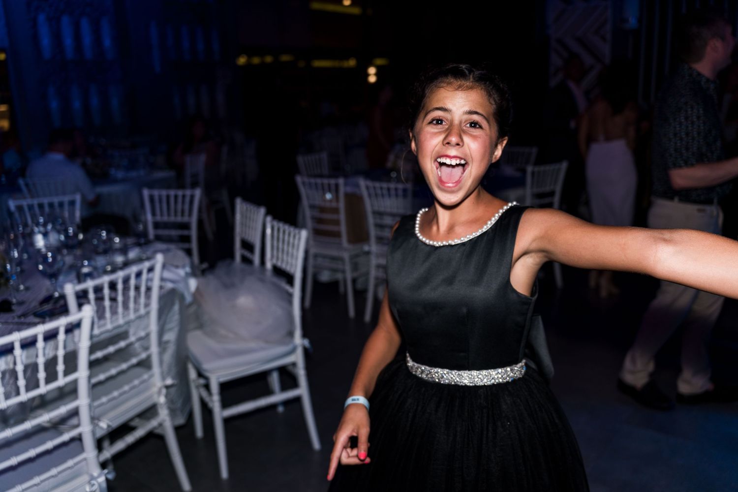Excited young girl in black dress with rhinestone belt laughs on wedding reception dance floor.