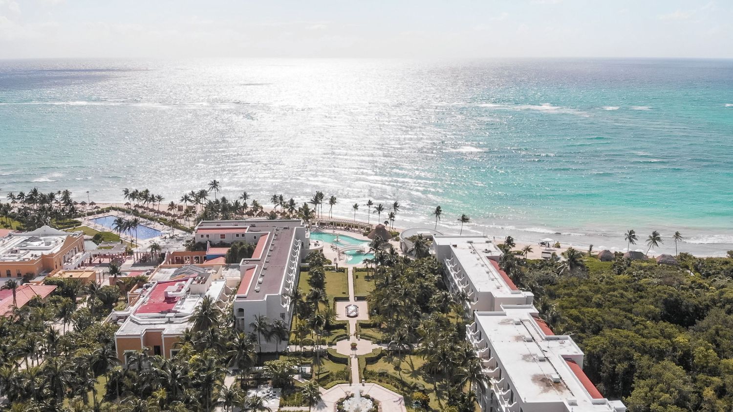 Aerial view of beachfront resort with turquoise Caribbean ocean, pool, and lush tropical gardens.