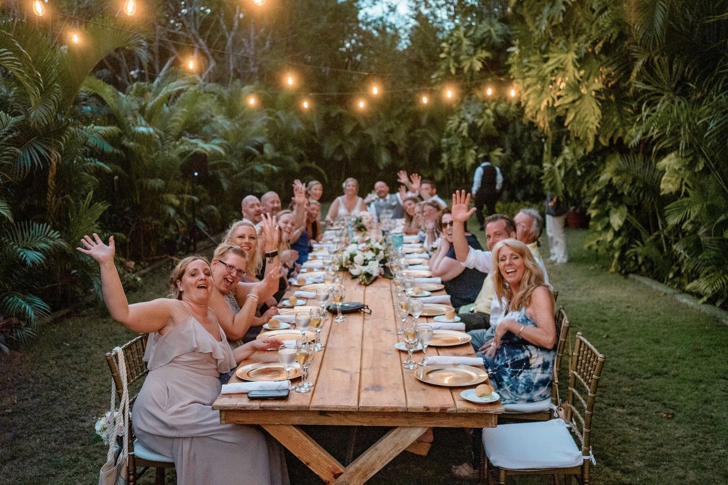 Guests celebrate at an outdoor wedding reception dinner under string lights surrounded by lush tropical greenery.