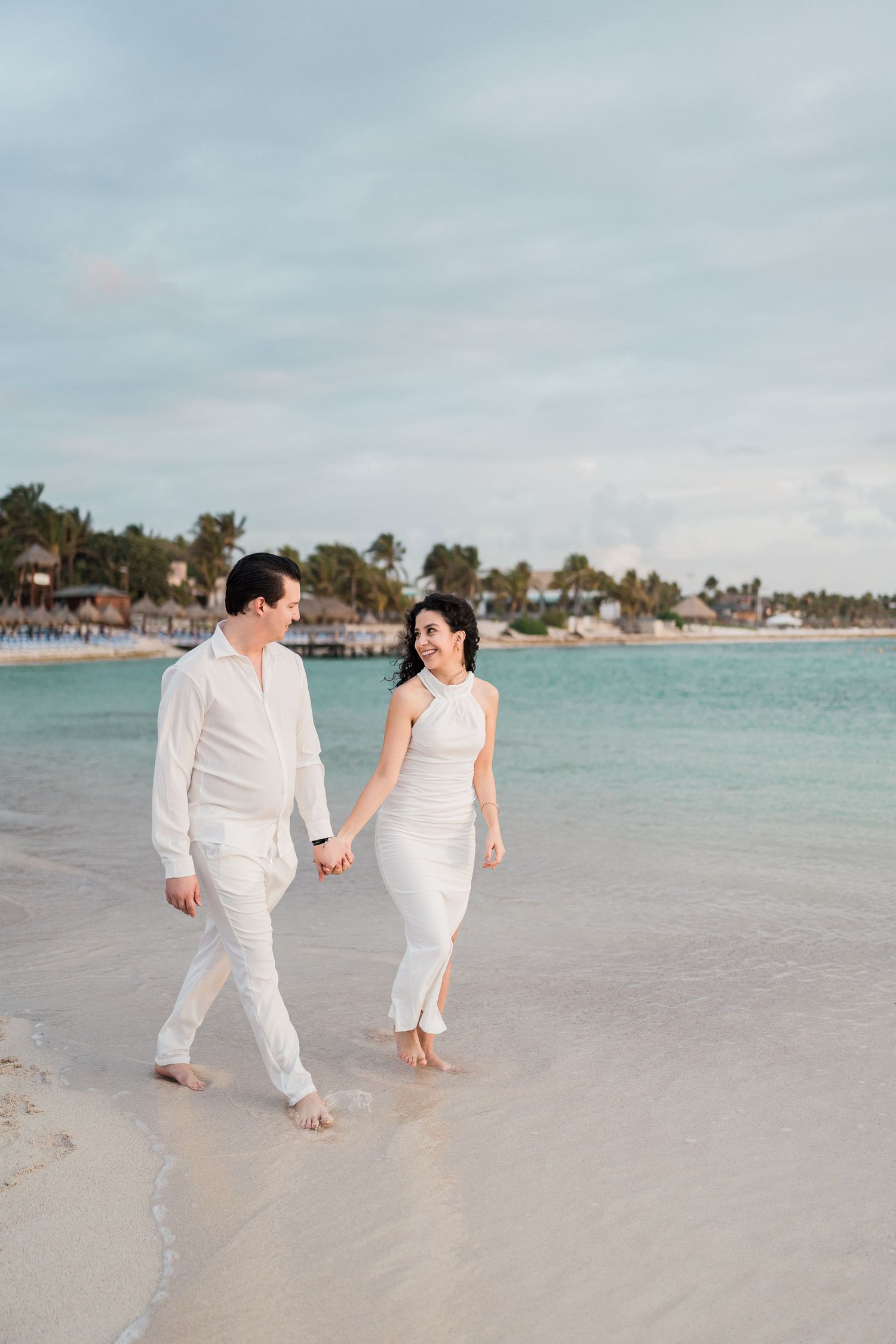 Couple in white outfits walking hand-in-hand on a tropical beach with turquoise water and palm trees.