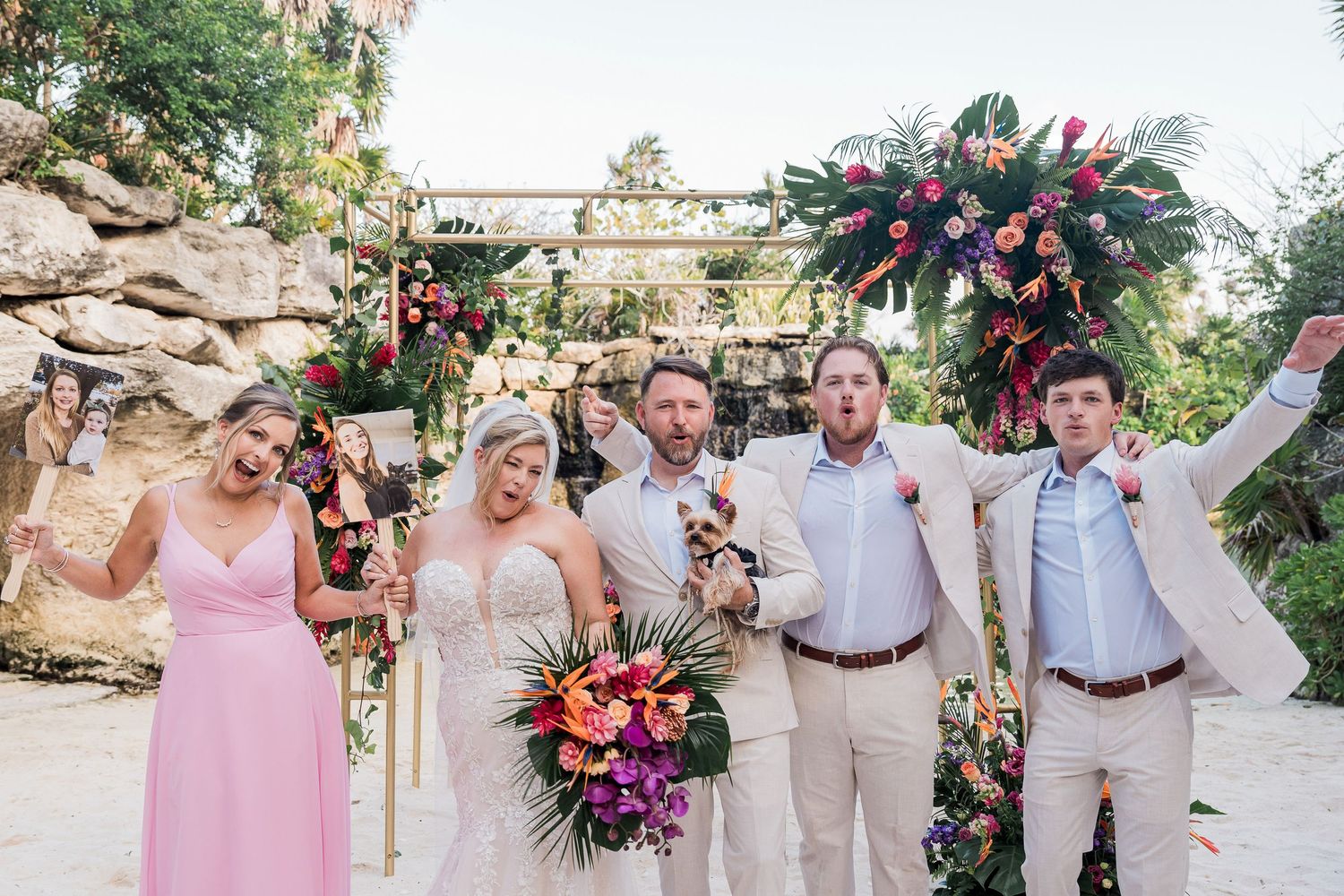 Joyful wedding party celebrating outdoors with tropical floral arch, bride holding colorful bouquet, groom and guests cheering.