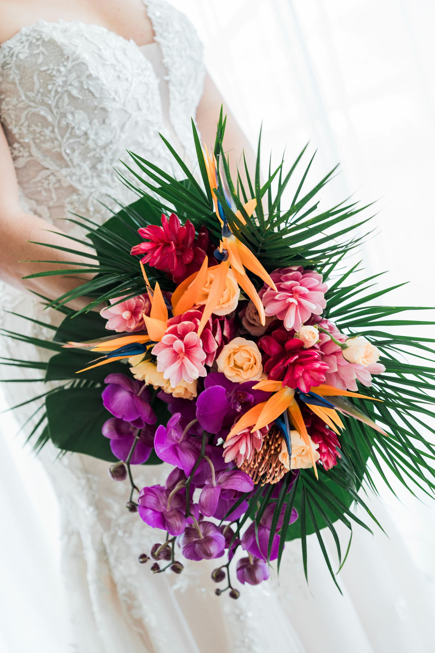 Bride holding a vibrant tropical wedding bouquet with orchids, birds of paradise, and palm leaves.