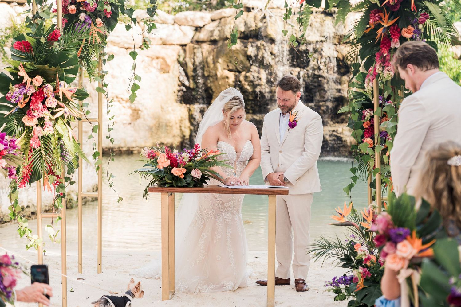 Couple signing marriage certificate at outdoor ceremony under colorful floral arch near waterfall.