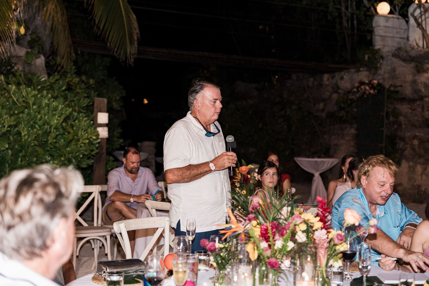 Man giving speech at outdoor tropical dinner party with colorful floral centerpieces and guests seated at tables.
