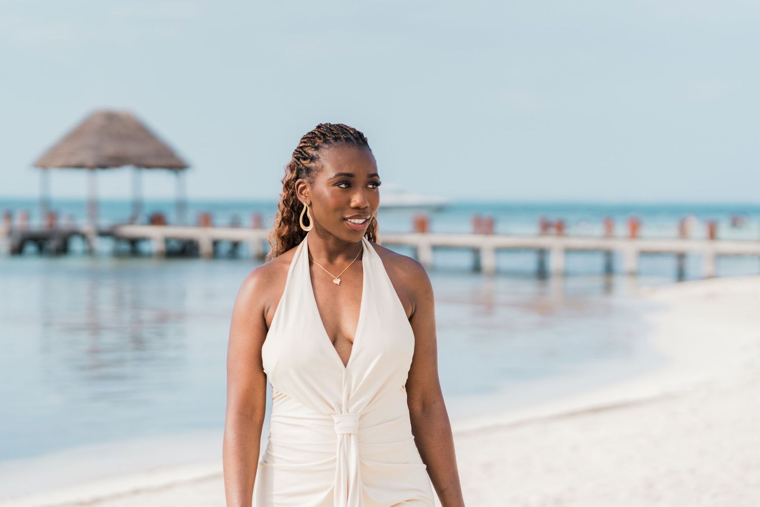 A woman in a white halter dress stands on a sandy beach with a pier and turquoise ocean in the background.