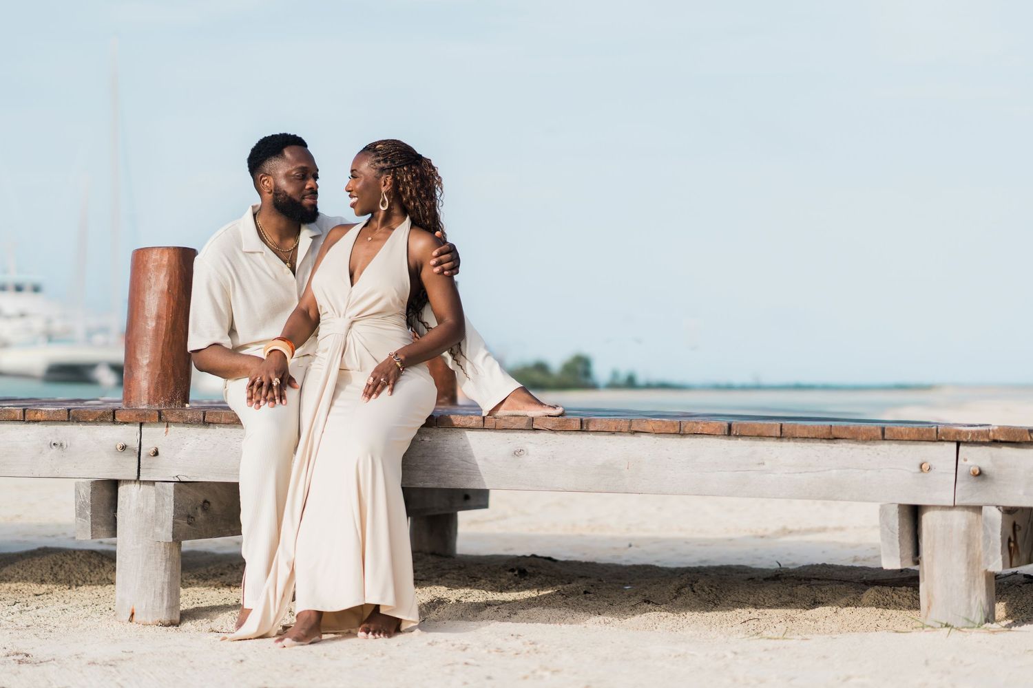Couple in cream attire sitting closely on a wooden bench near the beach.